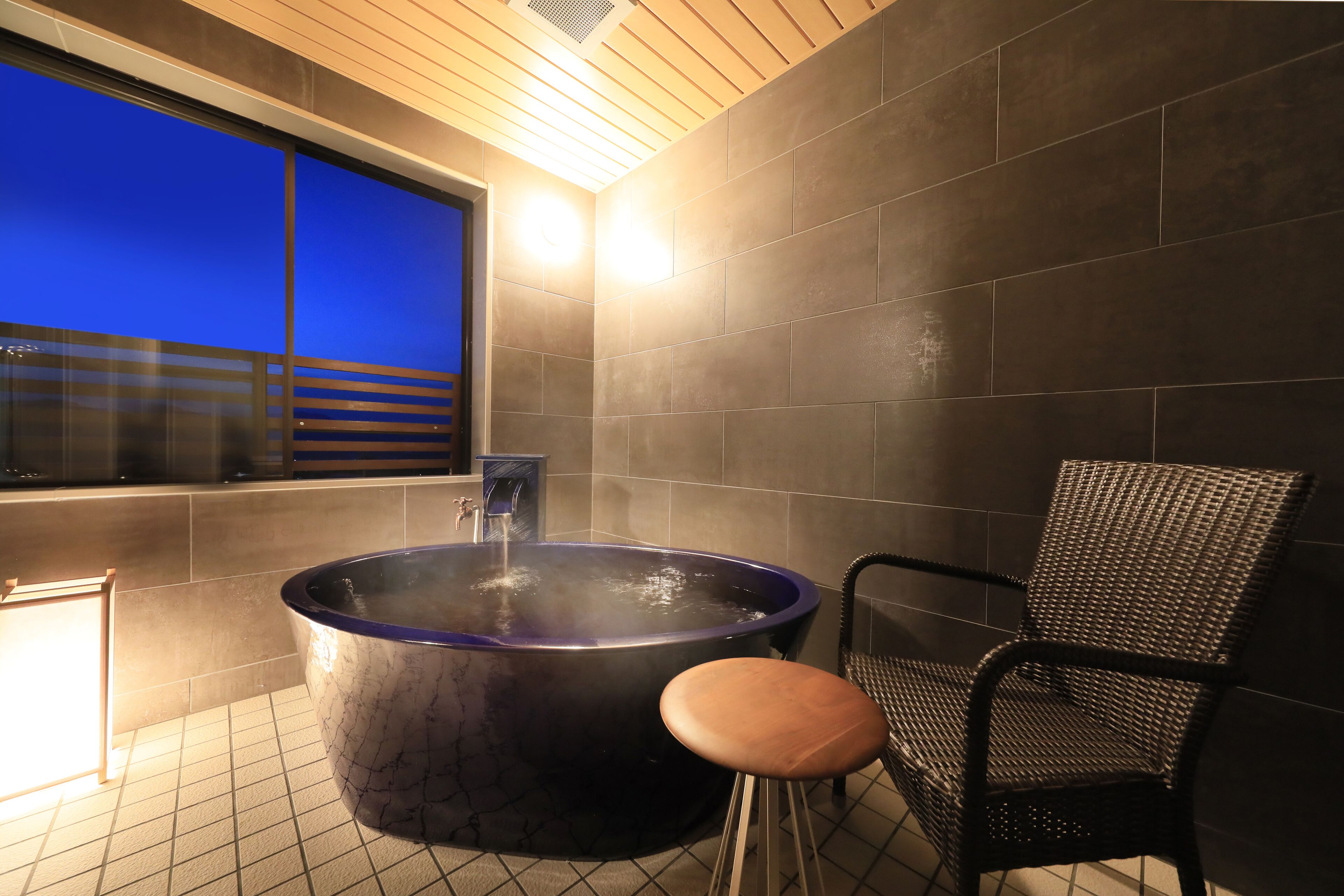 A modern bathroom with a large bathtub, a chair, and a window with a view of the cityscape.