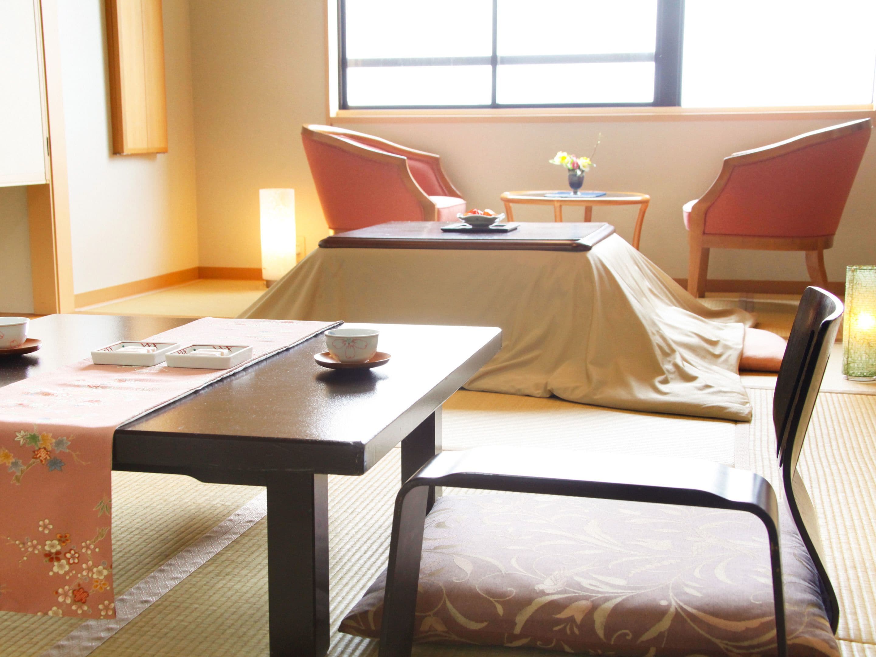Una habitación tradicional japonesa con suelo de tatami, una mesa baja con camino de mesa, dos sillas y una mesa pequeña con un jarrón de flores.
