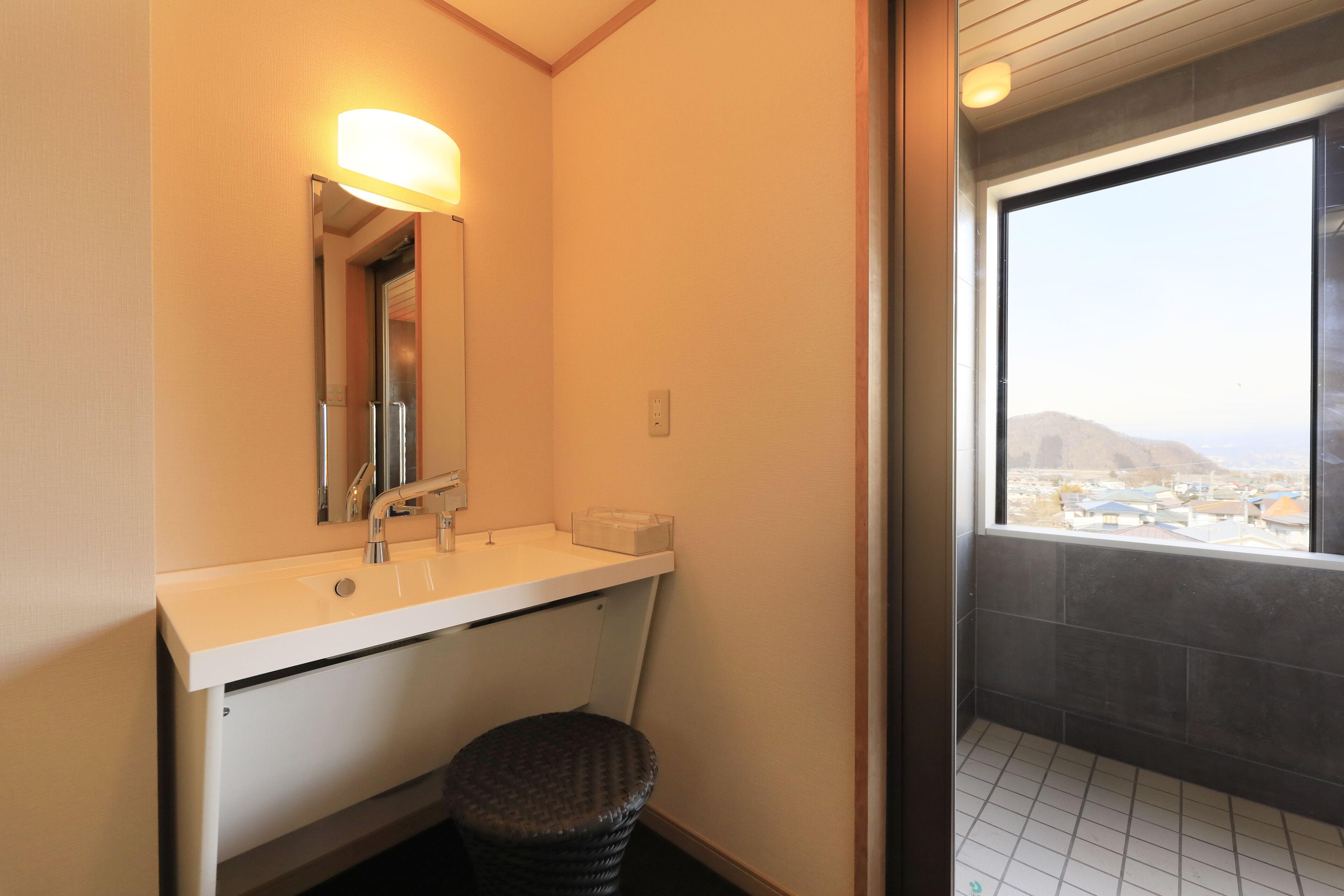 A bathroom with a white sink, a mirror, and a small stool. There is a window with a view of buildings and hills.