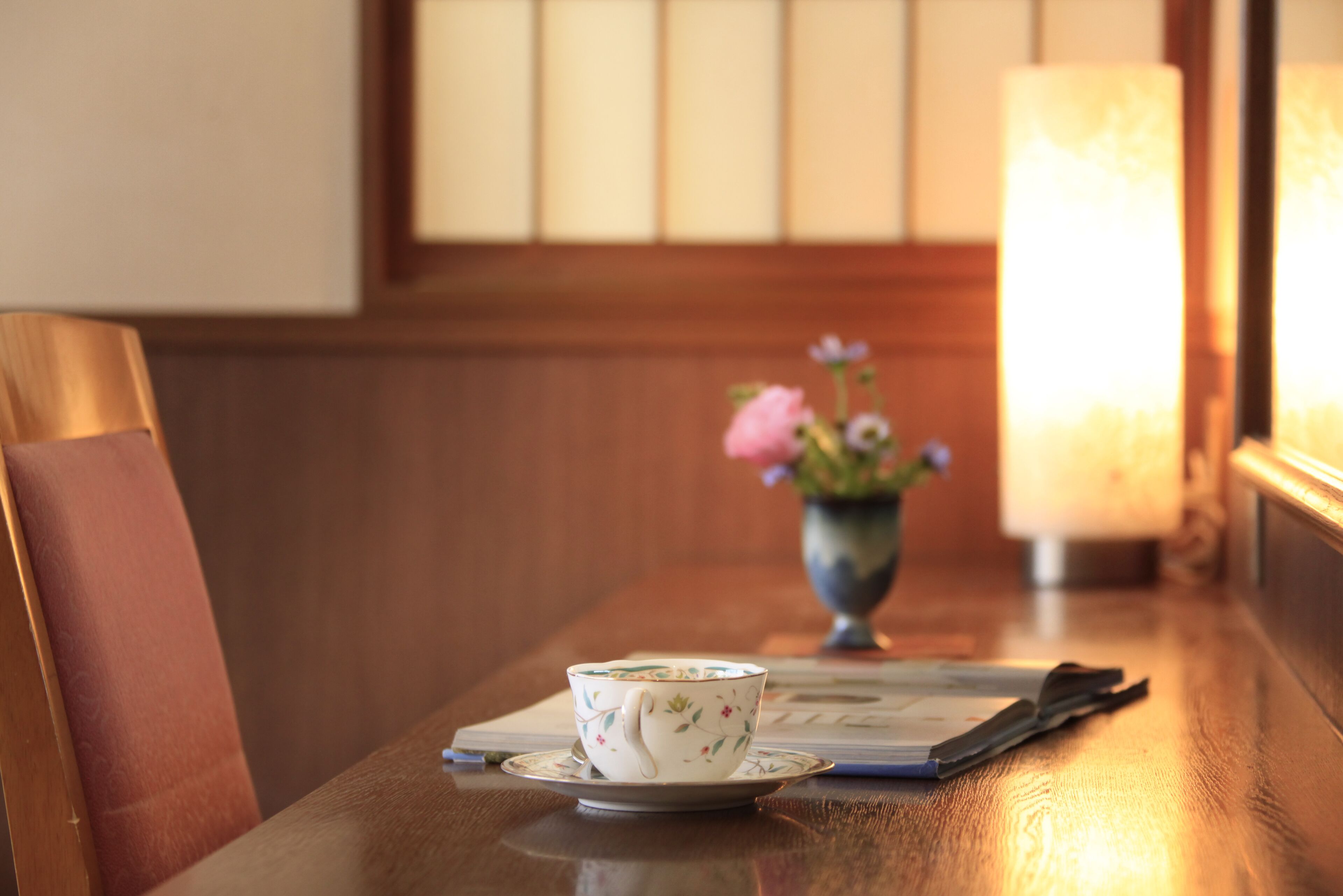 A wooden table with a cup and saucer, a vase with flowers, and a lit lamp.