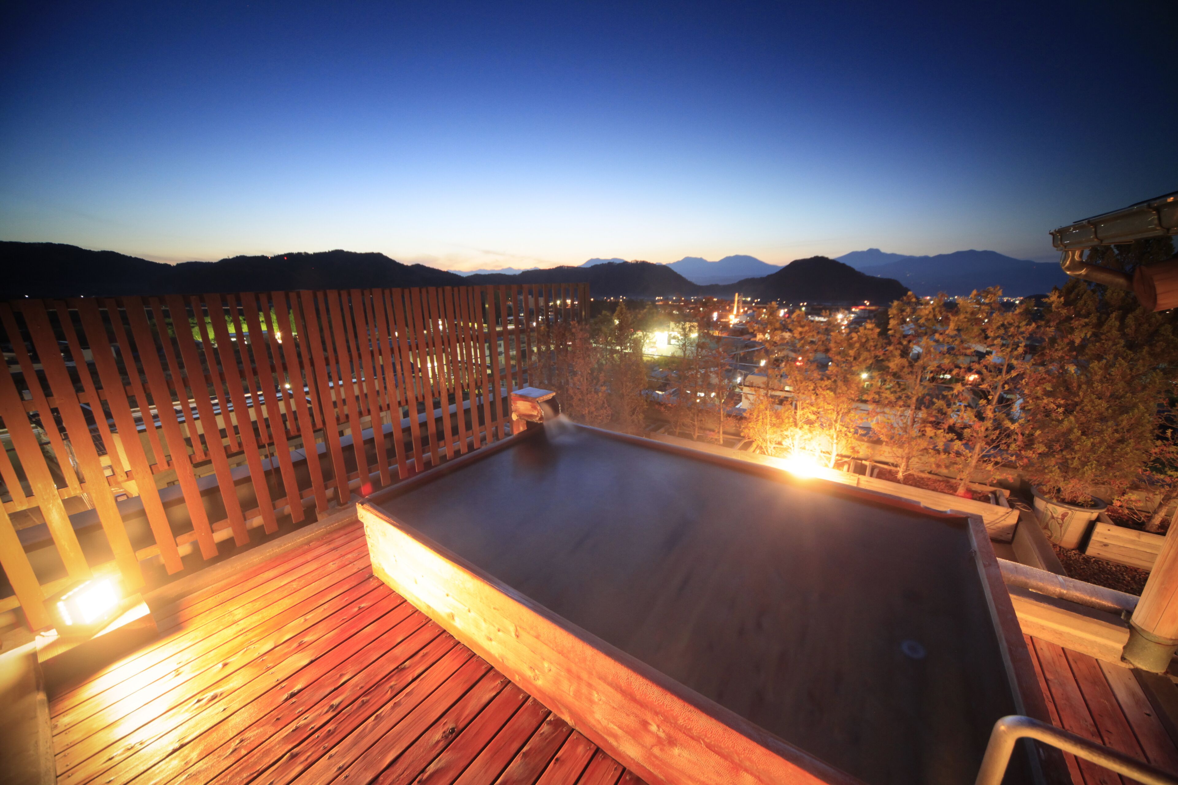 A wooden deck with a hot tub, surrounded by a wooden fence, overlooking a cityscape with mountains in the background.