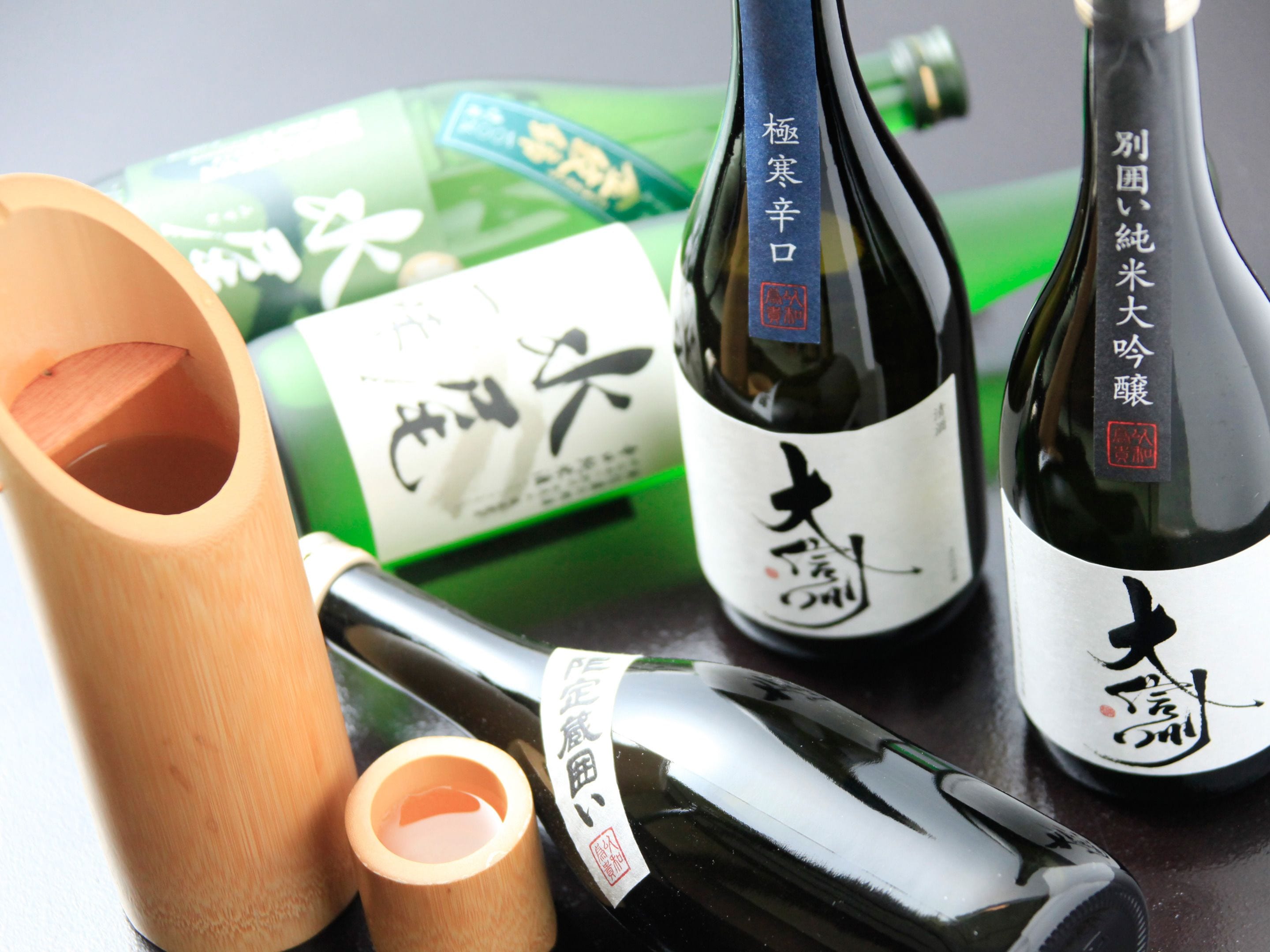 Three bottles of sake with Japanese labels, a wooden sake cup, and a green bottle with a label.