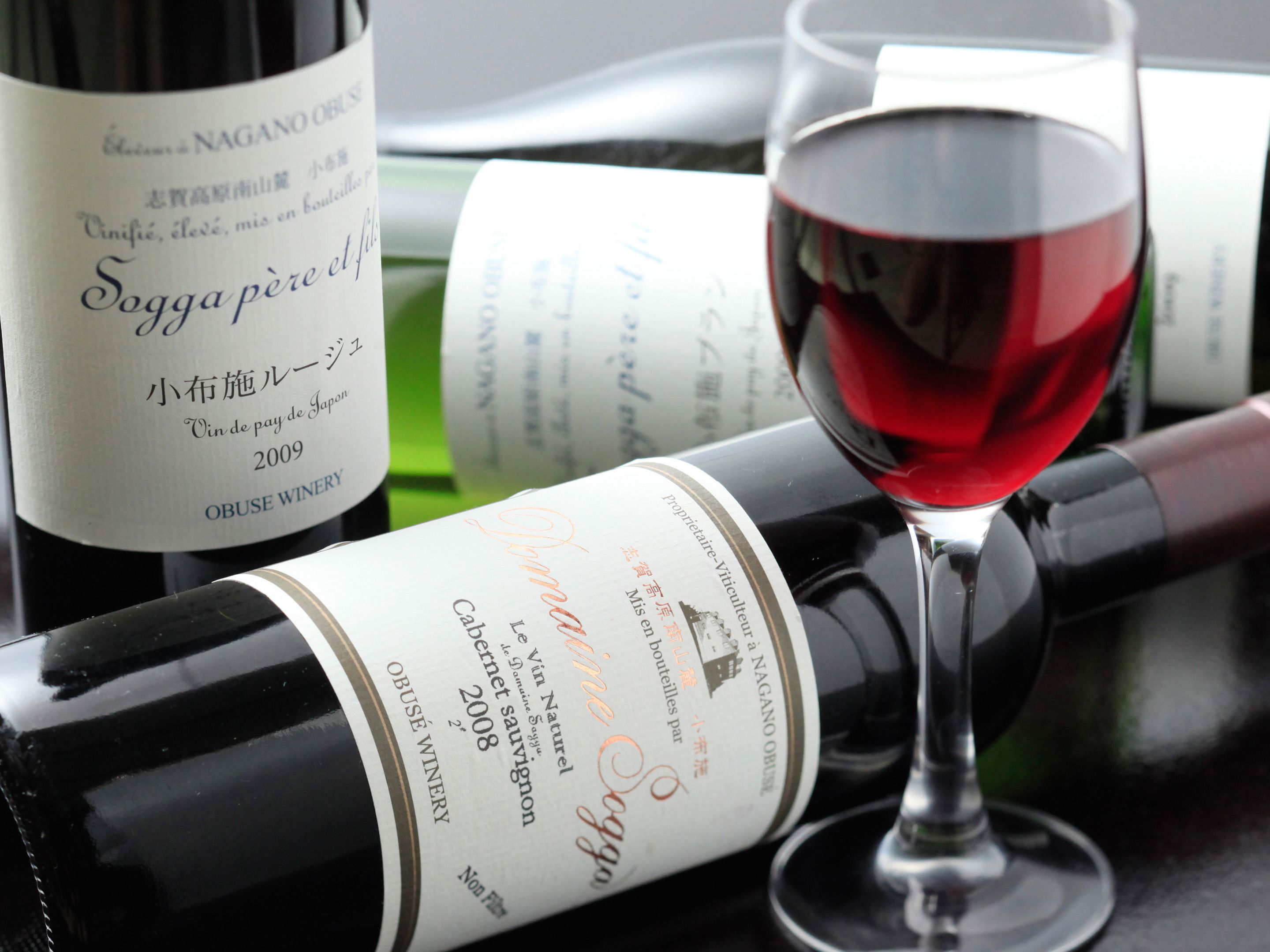 A wine bottle with a label, a wine glass with red wine, and other wine bottles in the background.
