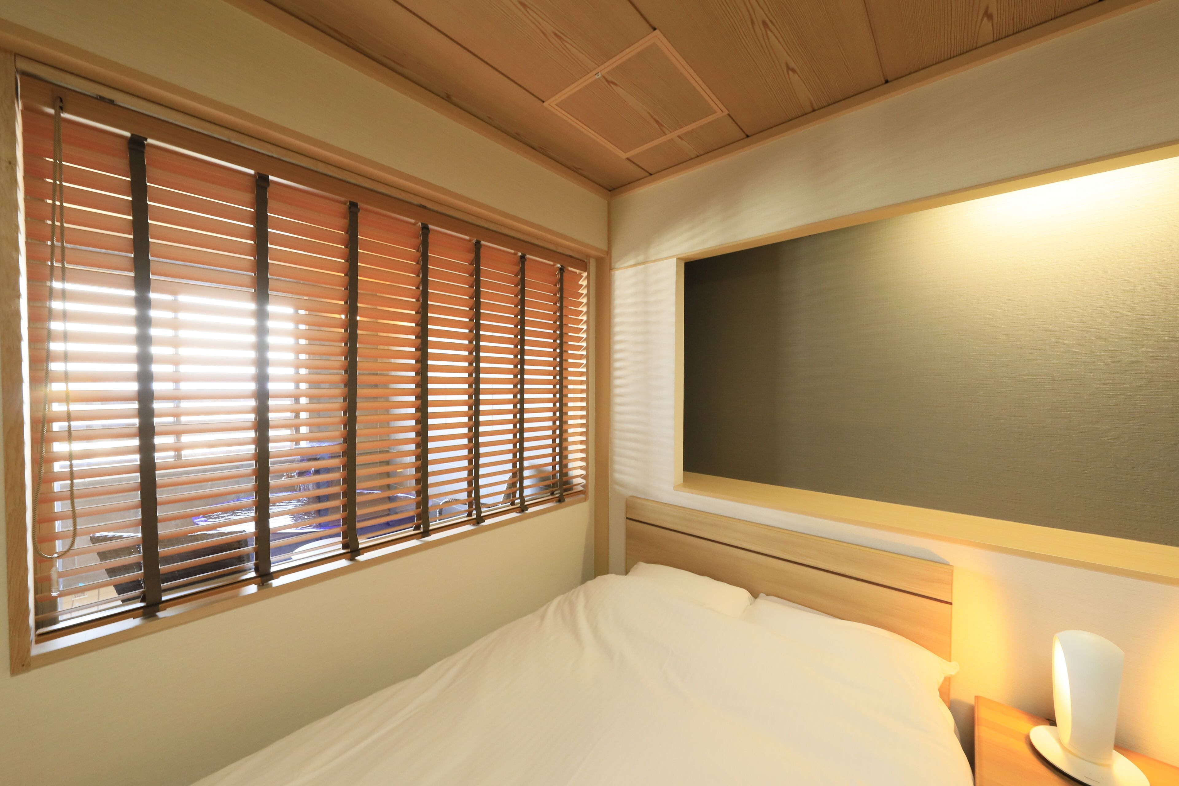 A bedroom with a bed, a nightstand, a wall-mounted lamp, and a window with blinds.
