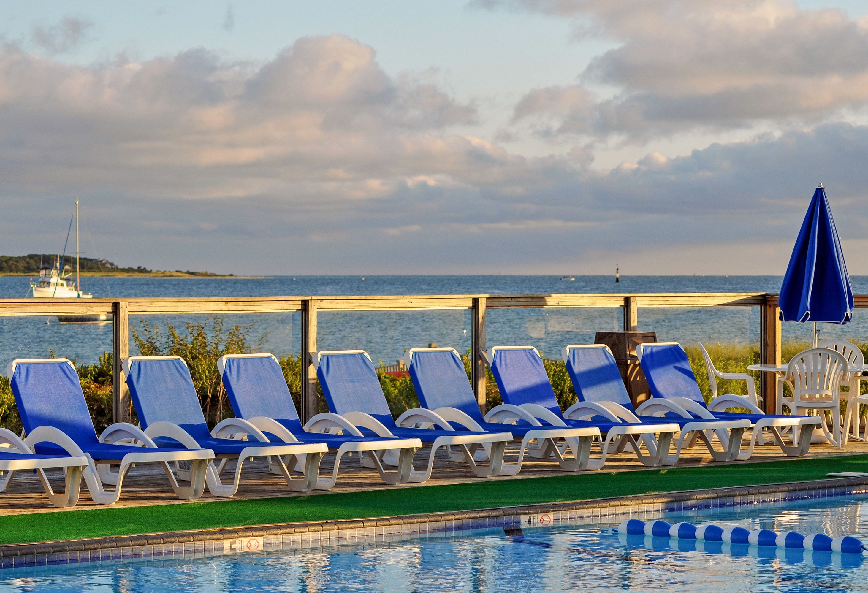 Un espace aménagé au bord de la piscine, avec des chaises longues et des parasols, offrant une vue sur une étendue d’eau et une île lointaine.