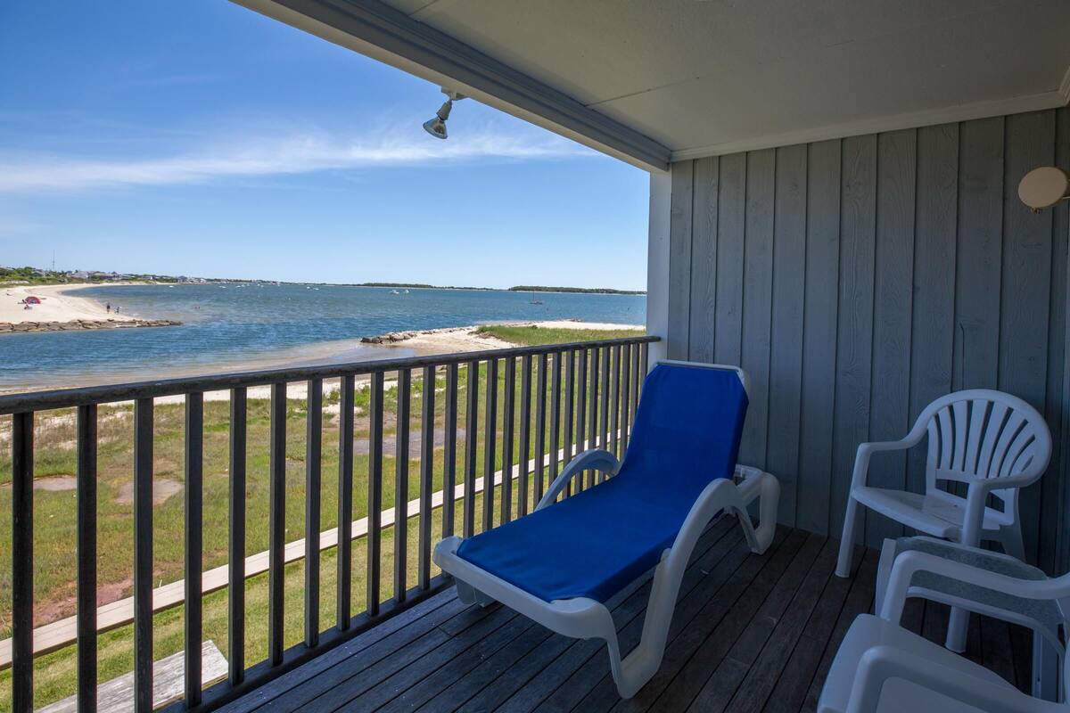 Ein überdachter Balkon mit Blick auf den Strand, ein blauer Liegestuhl und zwei weiße Stühle.