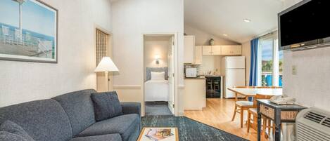 A compact hotel room with a grey sofa, a small wooden table, a TV, a kitchen area, and a dining table with chairs.