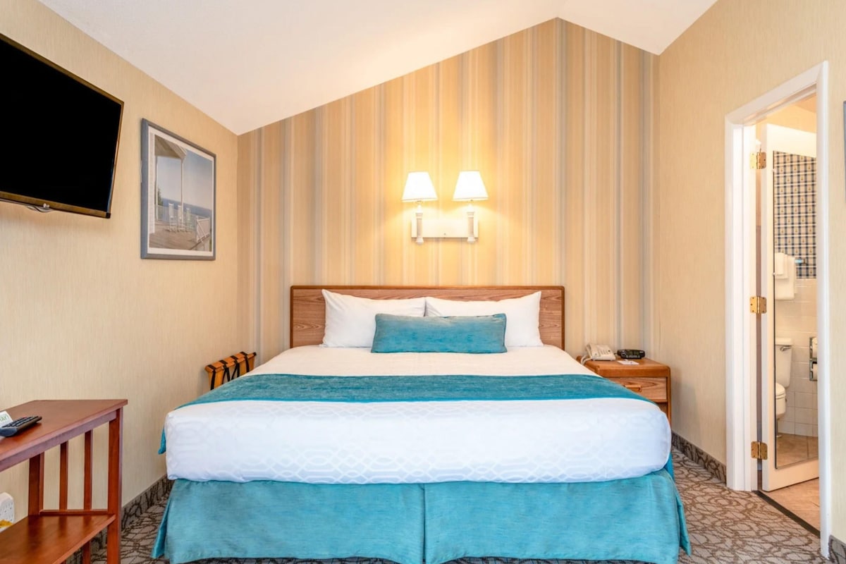 Habitación de hotel con una cama grande, un televisor de pantalla plana montado en la pared, una mesita de noche con lámpara y una puerta que da a un baño.