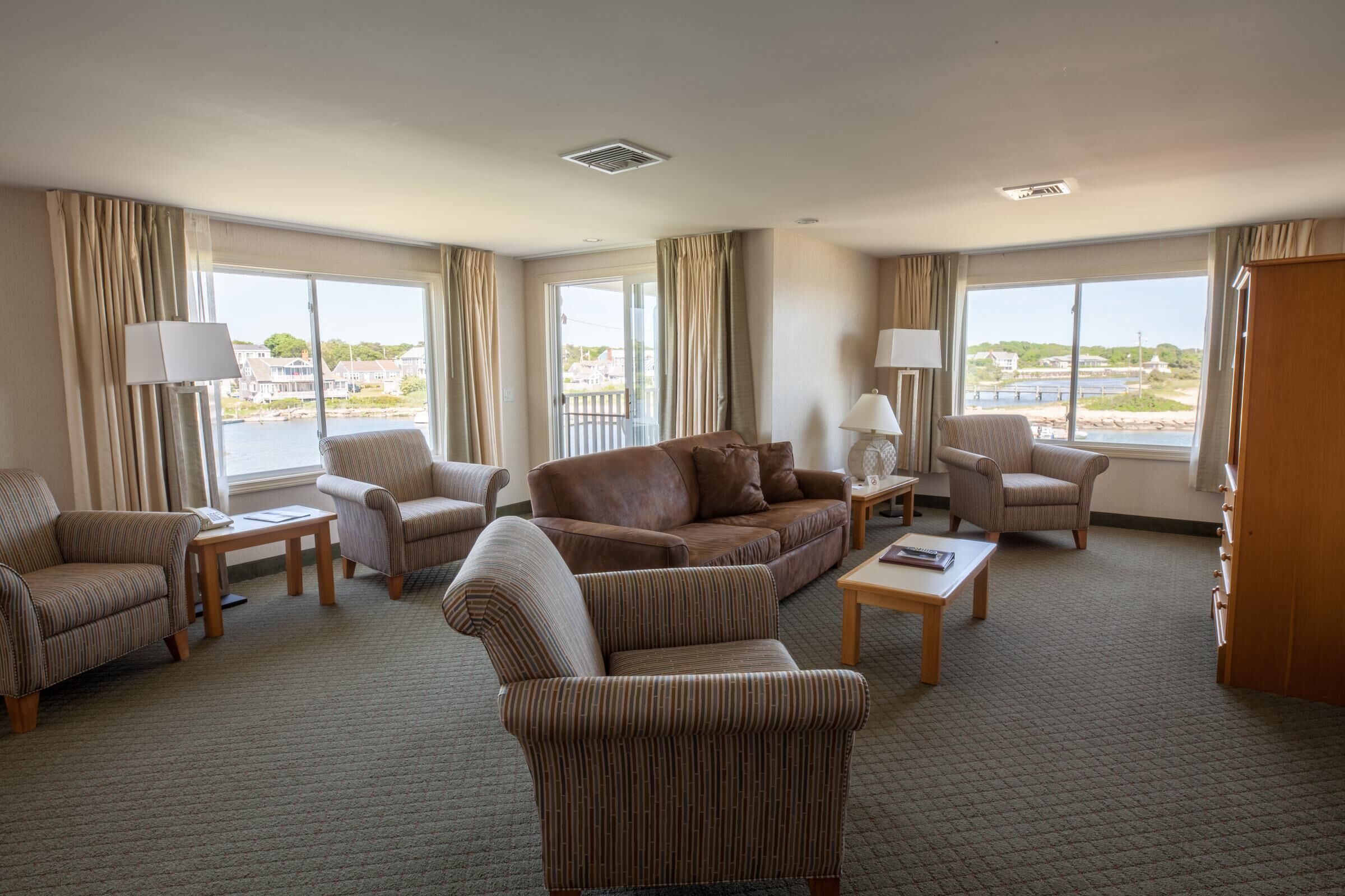 Un vaste salon d’hôtel comprenant un grand canapé, des fauteuils et une table basse. De grandes fenêtres offrent une vue sur l’eau et les bâtiments.