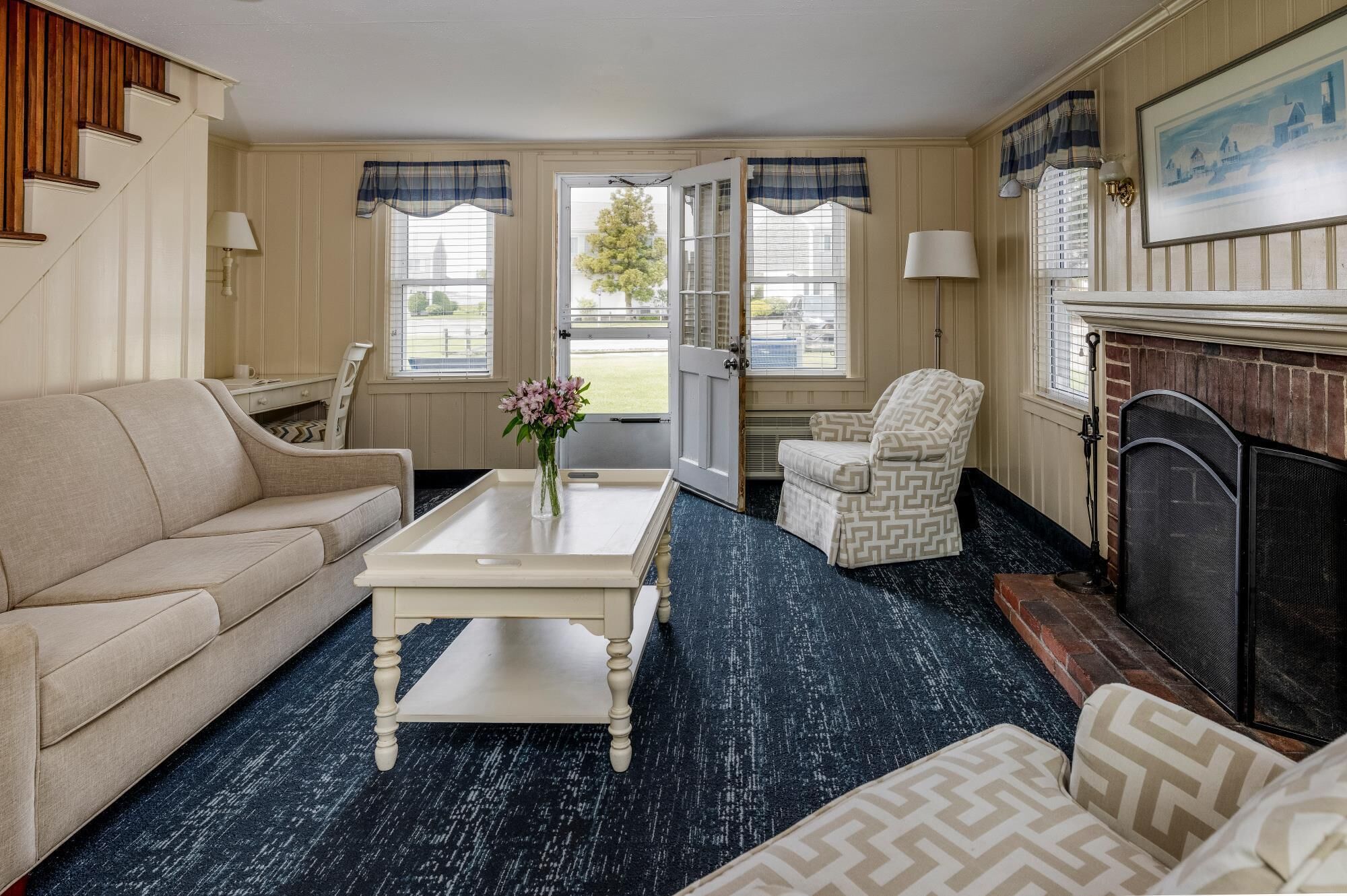 Un salon avec un canapé beige, un fauteuil à motifs, une table basse blanche ornée d’un vase de fleurs et une cheminée.