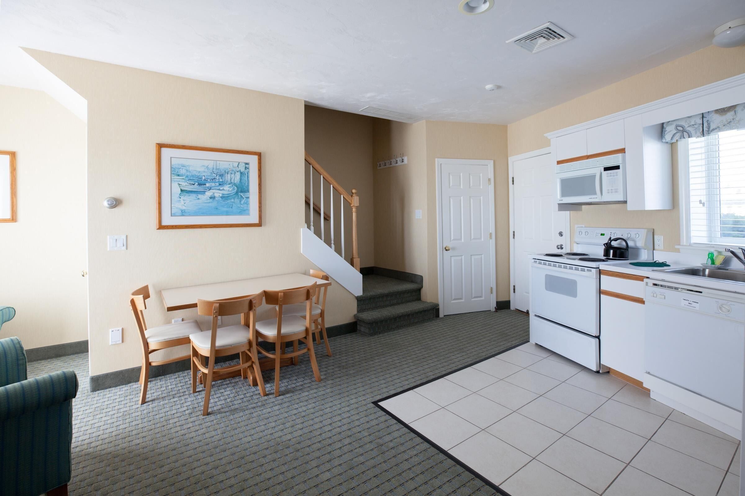 A kitchen with white appliances, a dining area with wooden chairs, and a staircase leading to another level.