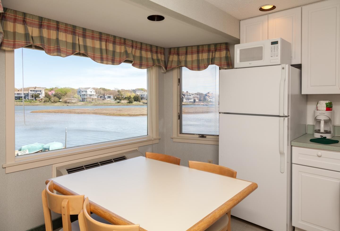A kitchen with a table and chairs, a refrigerator, a microwave, and a coffee maker. There are windows with a view of a body of water and houses.