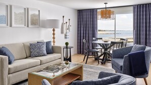 Ein modernes Wohnzimmer mit einer Couch, Sesseln, einem Couchtisch und einem Essbereich mit Stühlen und Tisch. An den Wänden befinden sich gerahmte Bilder, eine Lampe und durch große Fenster ist der Blick auf das Meer möglich.