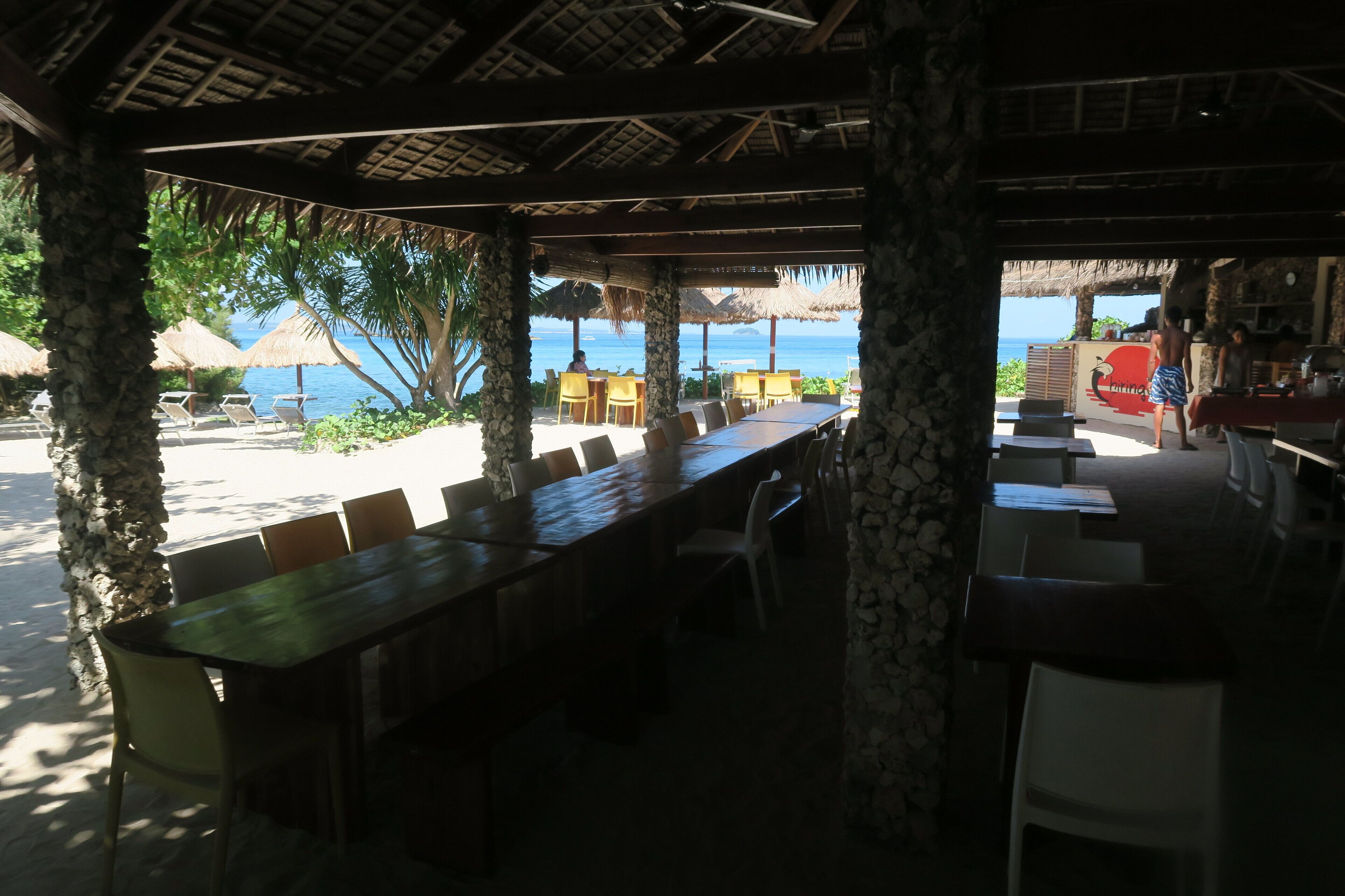 An outdoor dining area with a long wooden table and chairs, surrounded by stone pillars, overlooking a beach with umbrellas and chairs.