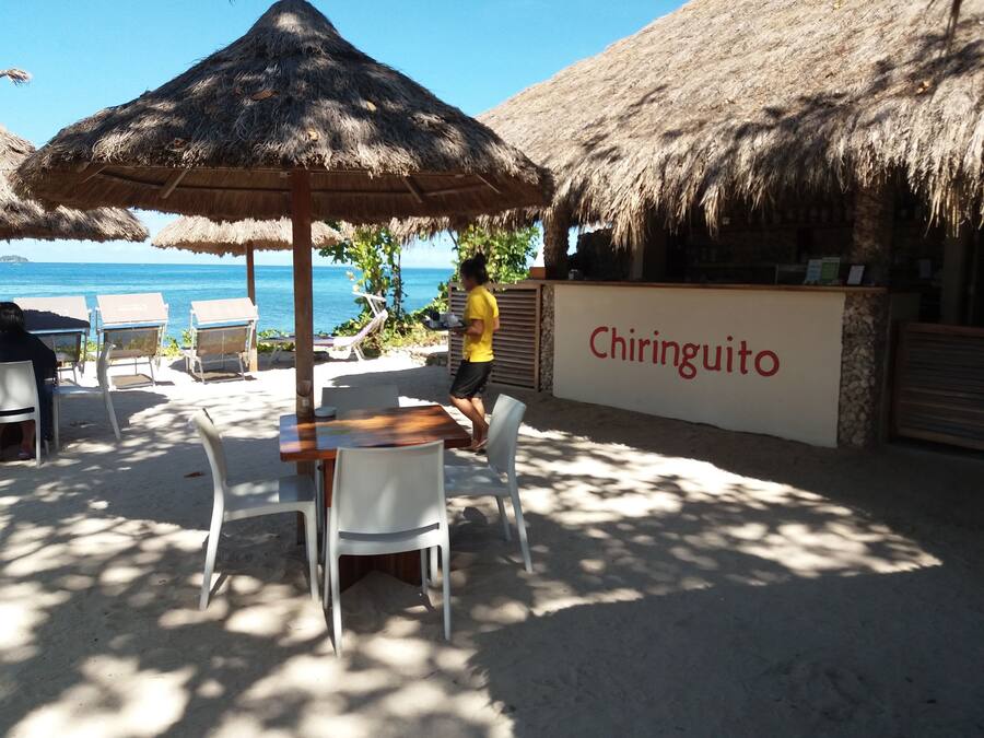 Un restaurante frente al mar llamado Chiringuito, con sombrillas de paja, sillas blancas y vistas al océano.
