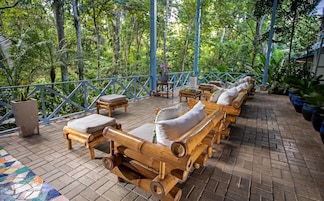 Un espacio de estar al aire libre cubierto, con sillones reclinables y mesas de madera, rodeado de abundante vegetación.