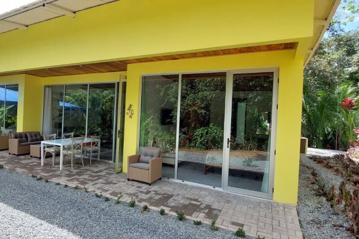 A yellow building with a covered patio area featuring outdoor furniture and a glass door leading to a garden.