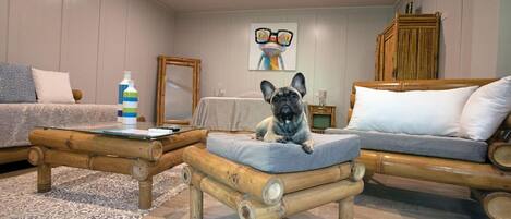 Un salón acogedor con un bulldog francés sobre un taburete acolchado, un sofá, una mesa de centro y un cuadro en la pared.