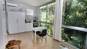 A compact kitchen with white cabinetry, a dining table with two chairs, and a large window overlooking a lush garden. - Hotel Plaza Yara (Quepos)