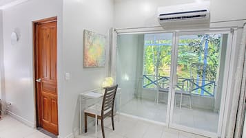 Une pièce avec une porte en bois, une petite table et une chaise, une porte vitrée donnant sur un balcon avec vue sur les arbres, et un appareil de climatisation fixé au-dessus de la porte.