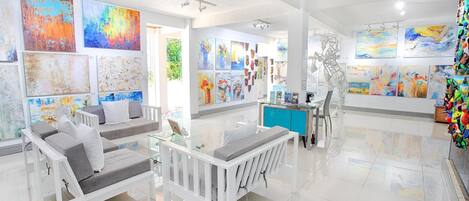 A modern art gallery with white furniture, including sofas and a glass table, and a variety of colorful paintings on the walls.