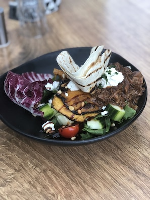 En svart tallrik med olika sorters mat, inklusive grillade grönsaker, en bit kött och en klick vit sås, placerad på ett träbord.