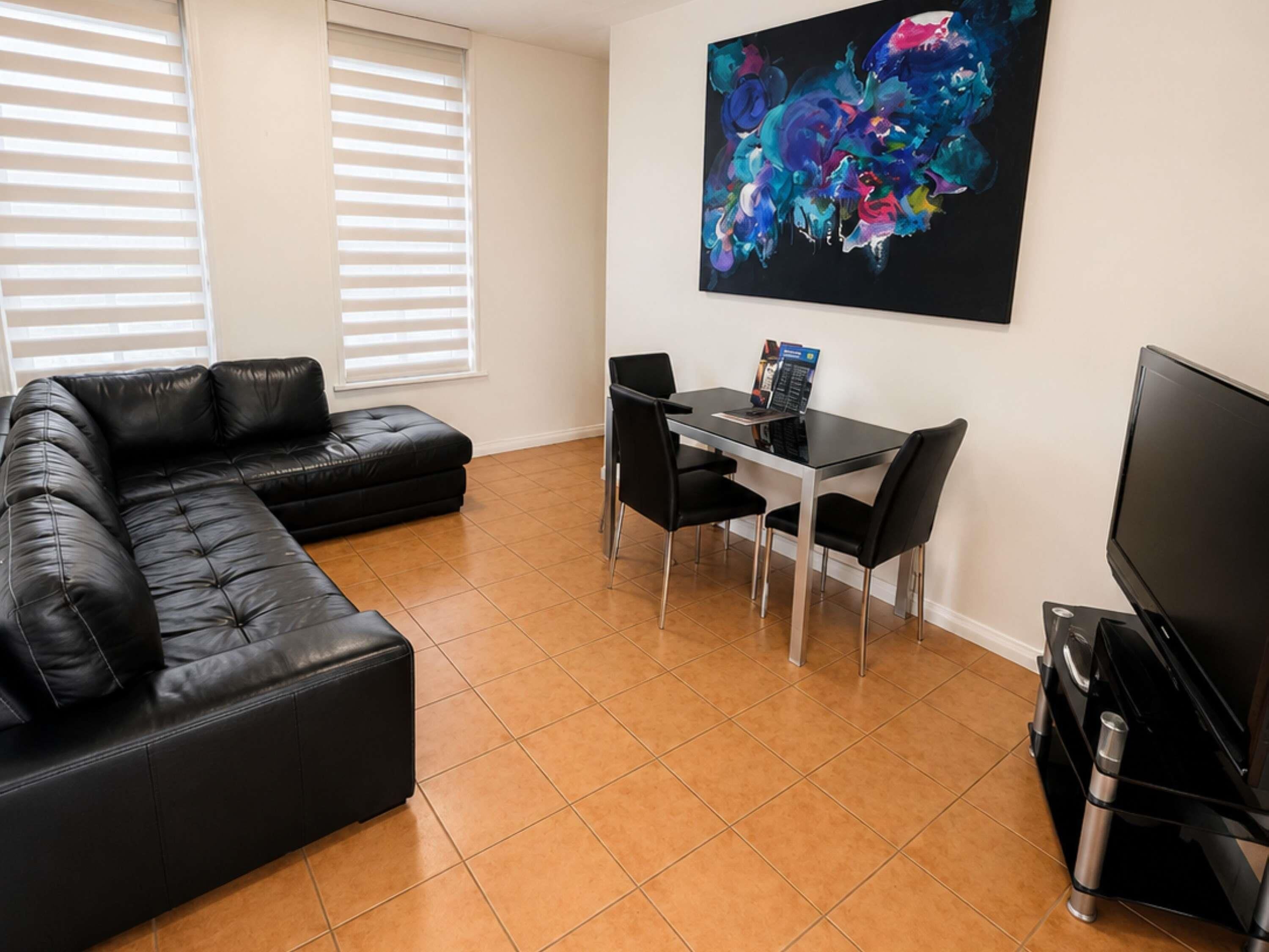 Una sala de estar con un sofá de cuero negro, una mesa de comedor con sillas, un televisor de pantalla plana y un cuadro abstracto grande en la pared.
