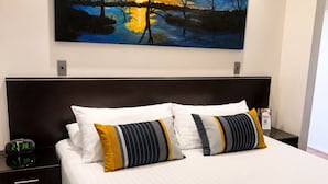 Een hotelkamer met een groot bed, twee nachtkastjes, een digitale klok en een schilderij van een zonsondergang boven het water.