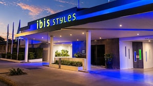 Een moderne hotelentree met een bord waarop 'ibis Styles' staat, een overdekte wachtruimte en een heldere lucht.