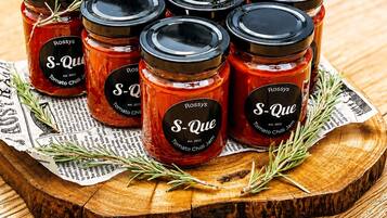 Six jars of S-Que Tomato Chilli Jam on a wooden surface with newspaper and rosemary sprigs.