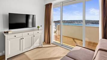 Una habitación moderna con una televisión de pantalla plana sobre un mueble blanco, un amplio ventanal corredero a un balcón con vistas al mar y edificios, y un sofá color crema.