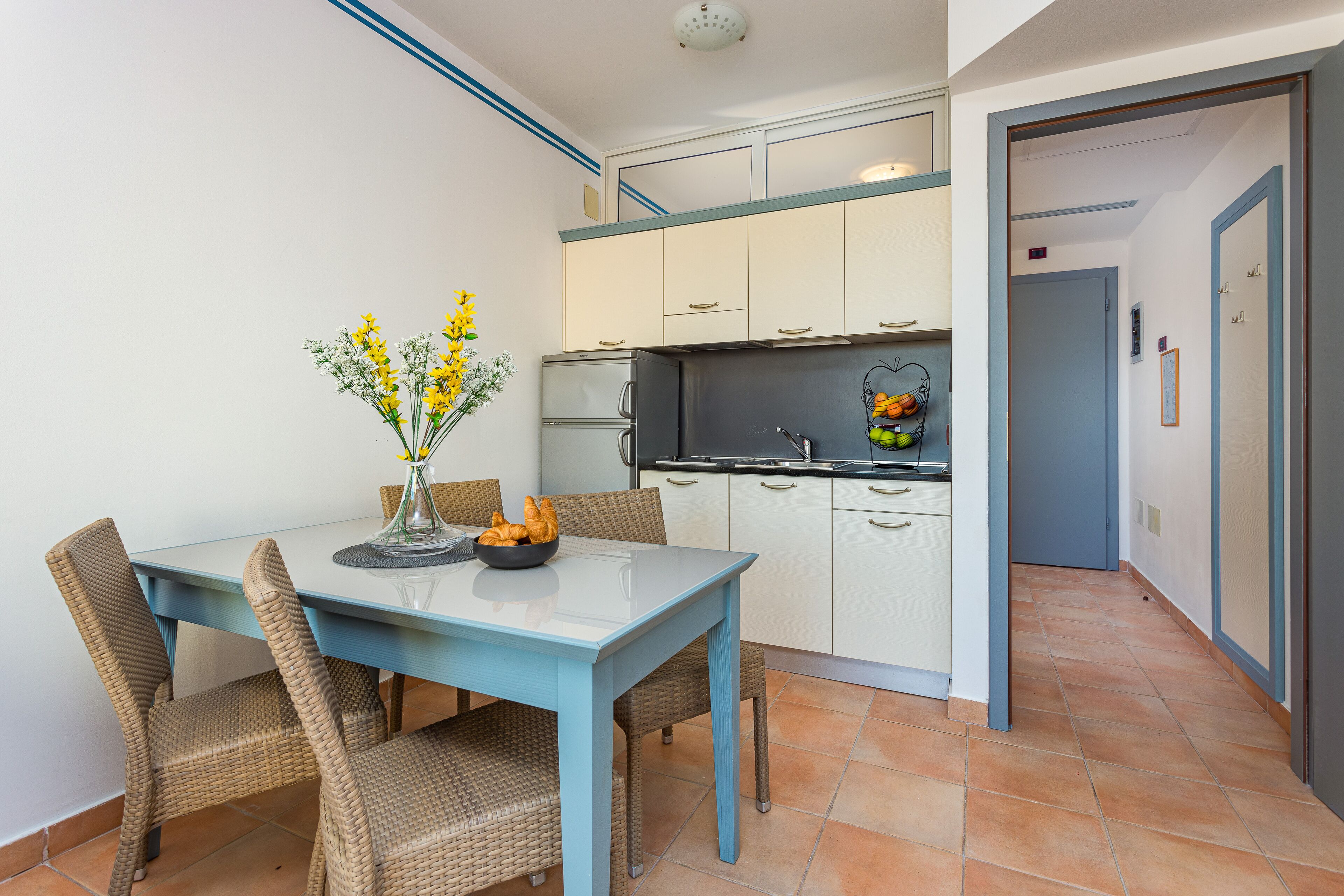 A modern kitchen with white cabinets, a blue table, wicker chairs, and a vase with flowers.