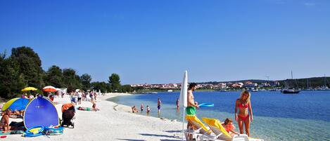 En strand med kristallklart blått vatten, folk som solar och båtar i fjärran.