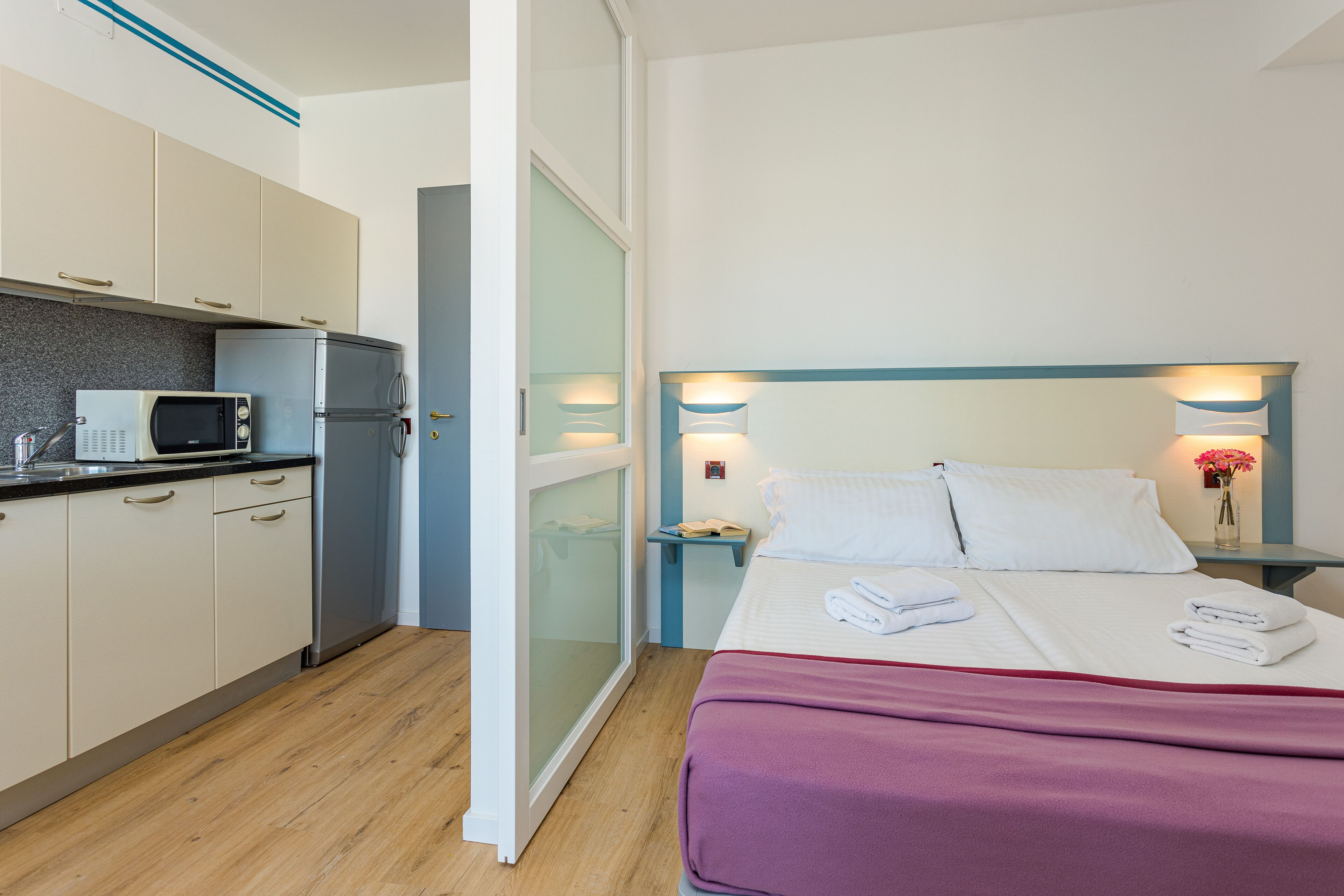 A compact hotel room with a kitchenette, a bed with a purple bedspread, and bedside lamps.