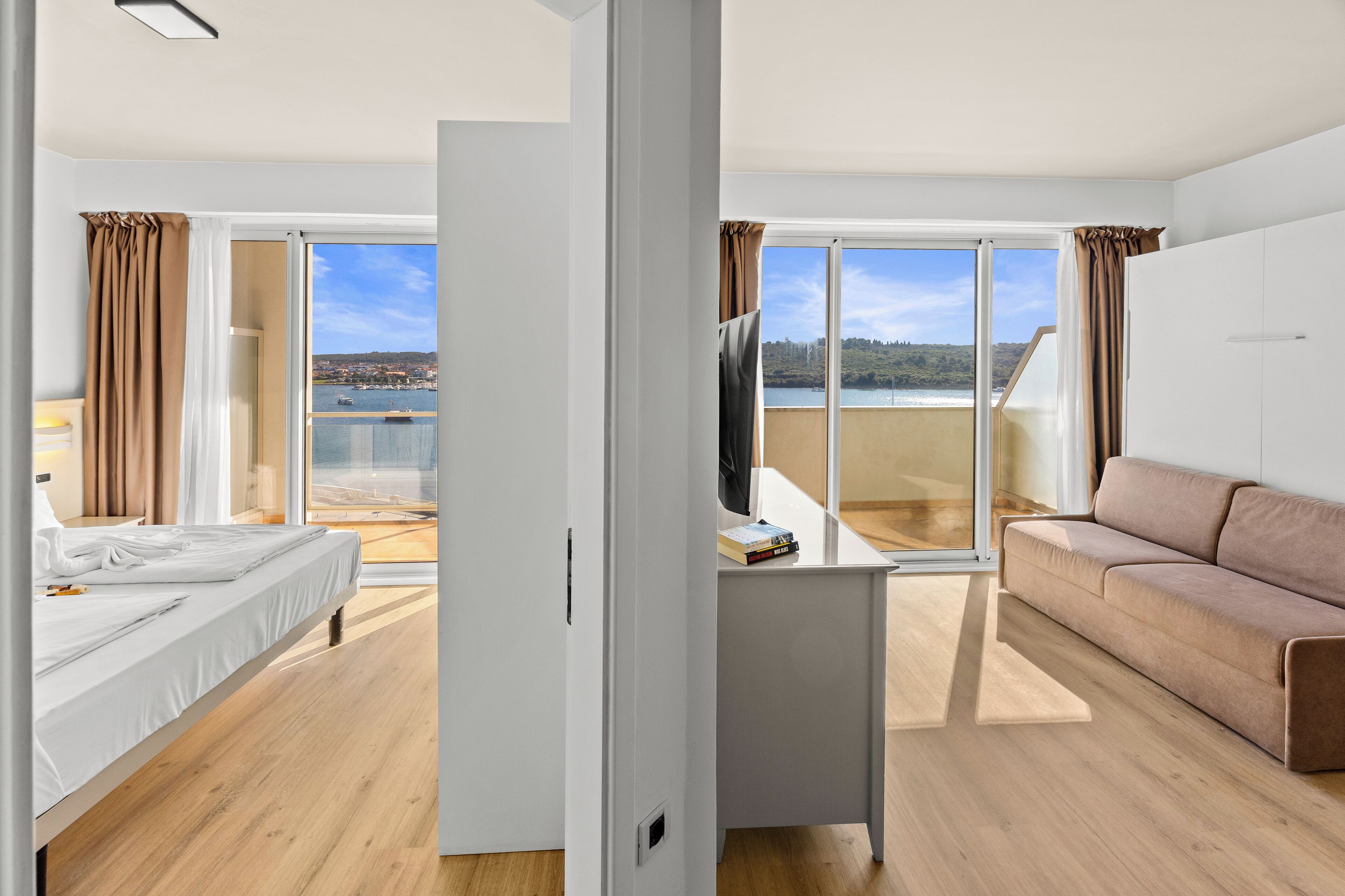 Une chambre d’hôtel moderne équipée d’un lit, d’un canapé, d’un téléviseur posé sur un meuble et d’un balcon offrant une vue sur l’eau et les collines.