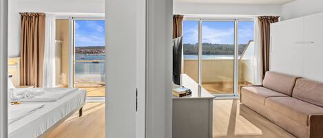 Une chambre d’hôtel moderne équipée d’un lit, d’un canapé, d’un téléviseur posé sur un meuble et d’un balcon offrant une vue sur l’eau et les collines.
