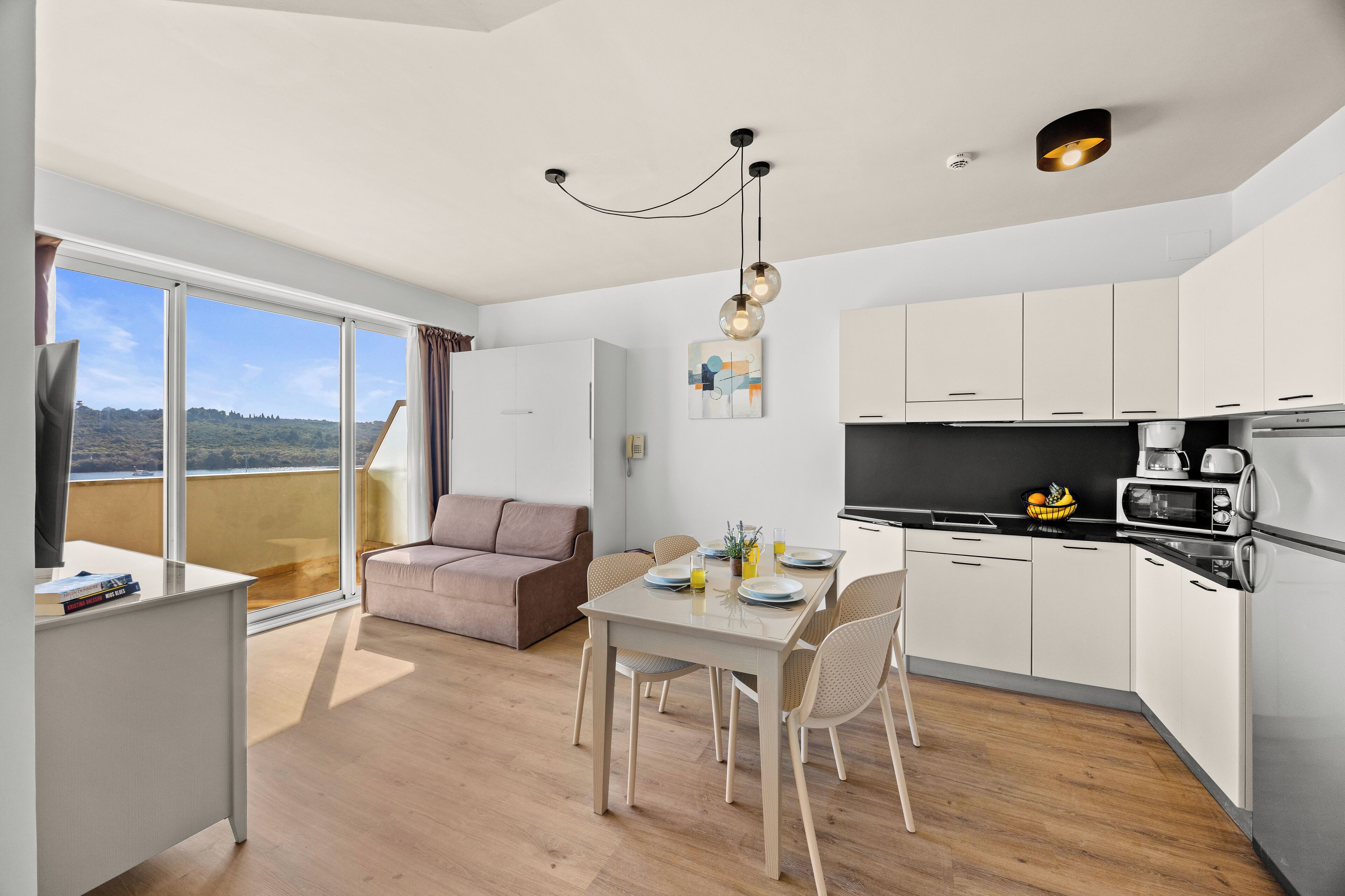 Une cuisine moderne avec des meubles blancs, un coin repas comprenant une table et des chaises, un canapé et une grande fenêtre donnant sur l’extérieur.