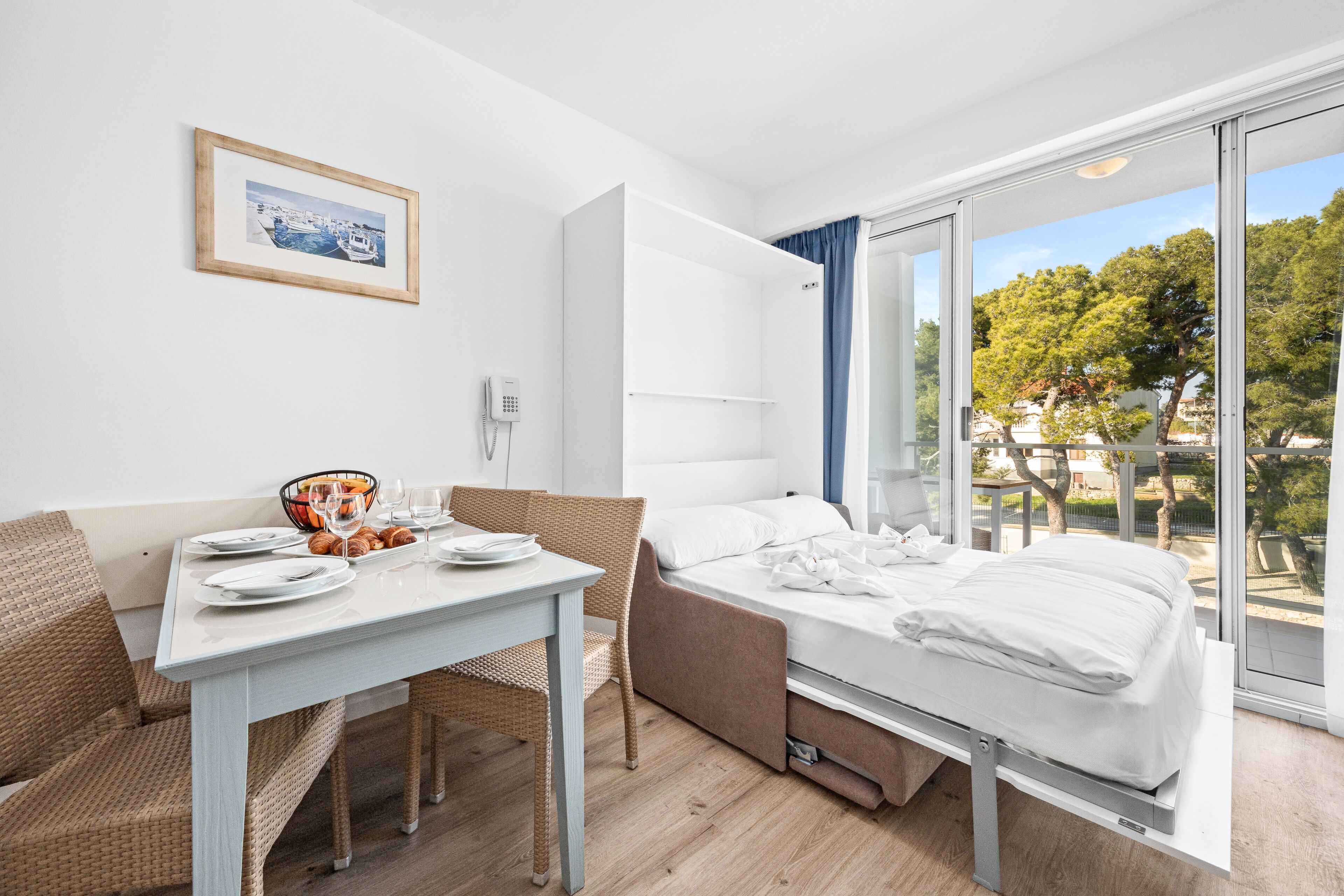 Una habitación de hotel moderna con cama, una mesa de comedor con sillas, una pequeña cocina y un balcón con vistas a árboles y edificios.