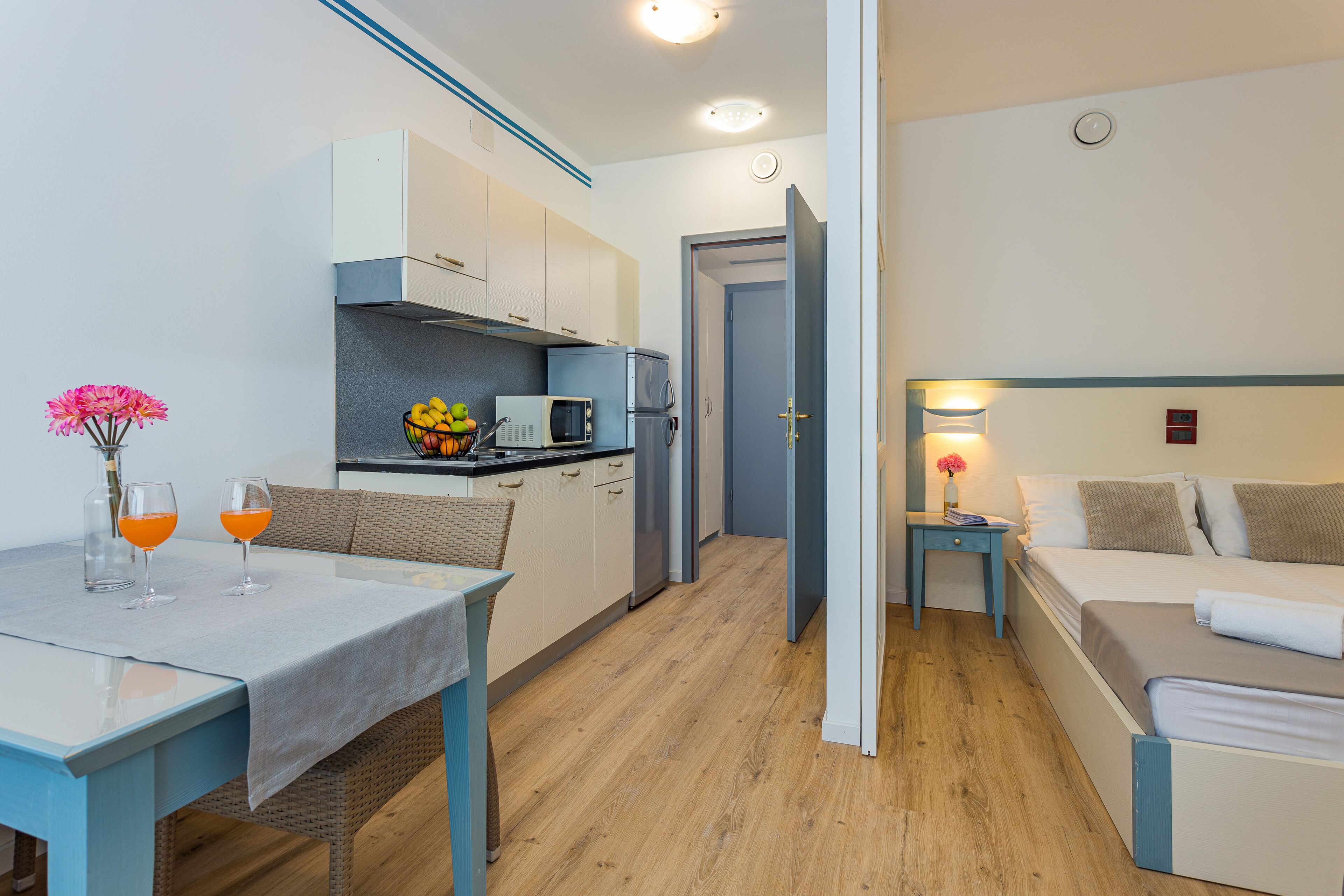 A compact hotel room with a kitchenette, a dining area with a table and two glasses of orange juice, and a bed with a nightstand.