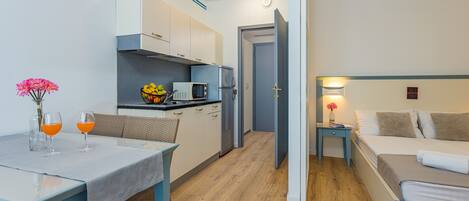 A compact hotel room with a kitchenette, a dining area with a table and two glasses of orange juice, and a bed with a nightstand.