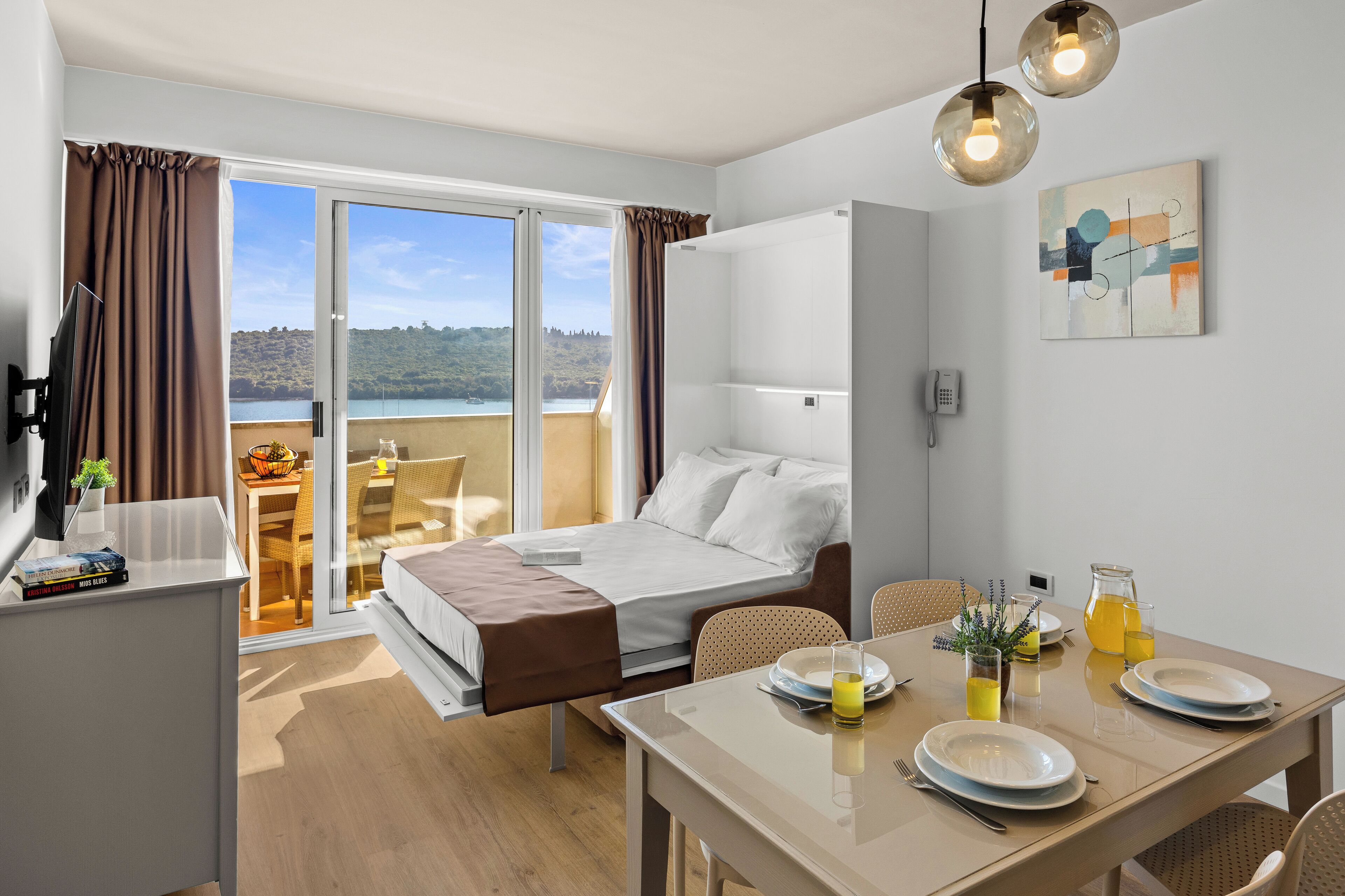 Habitación de hotel con cama, una mesa de comedor con alimentos para el desayuno, vista al exterior y un cuadro en la pared.