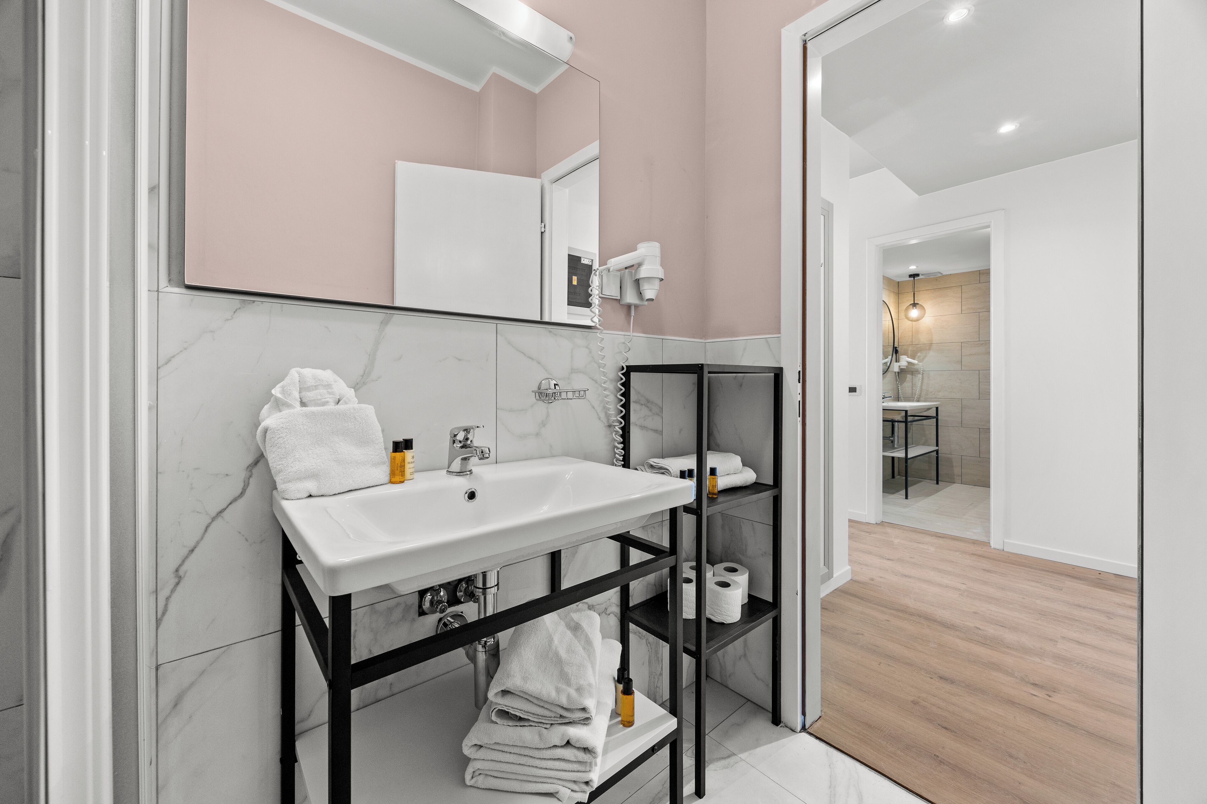 Une salle de bain moderne avec un lavabo blanc, des murs en marbre, une étagère en métal noir et un plancher en bois.