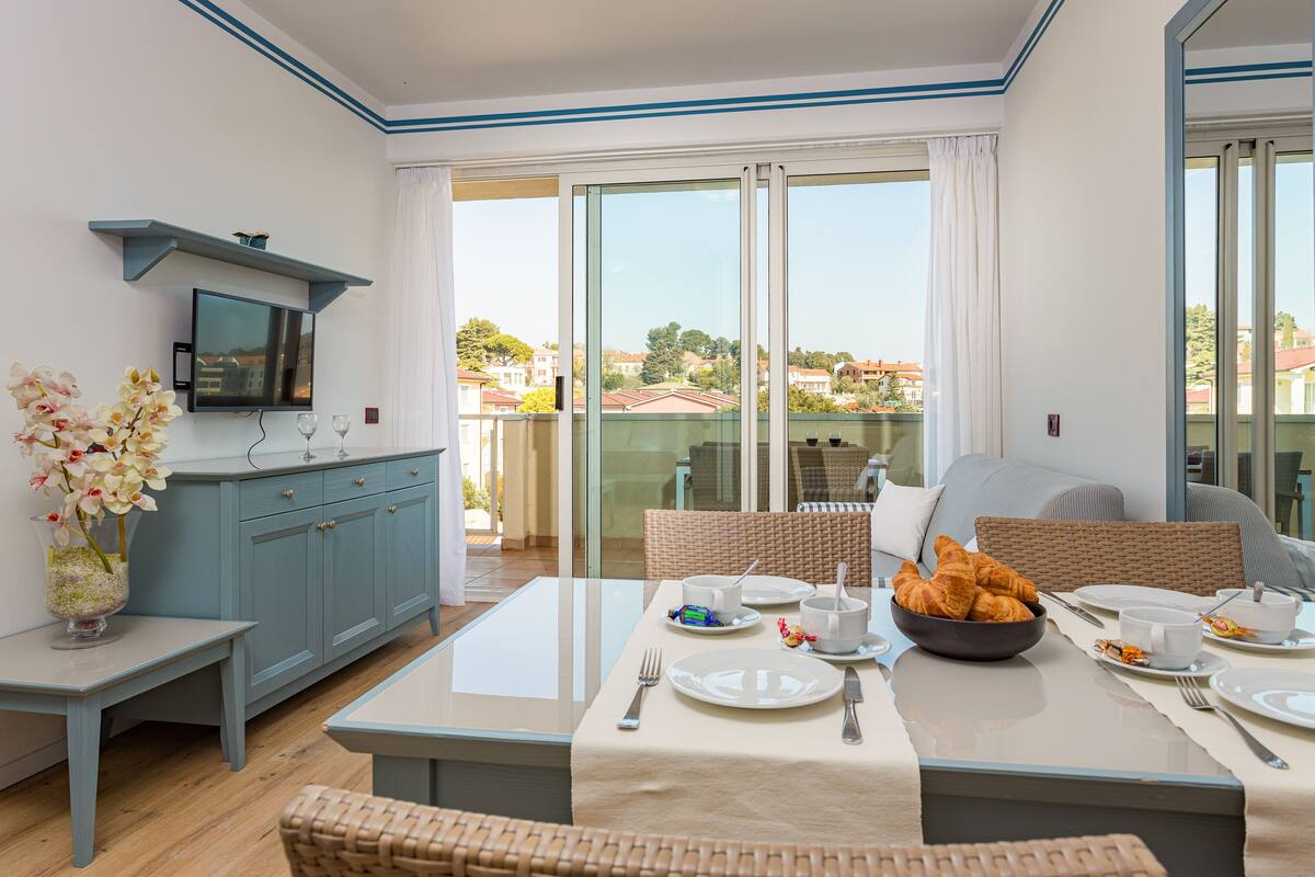 Un comedor moderno con una mesa dispuesta para el desayuno, un televisor sobre la encimera y vistas al vecindario a través de amplios ventanales.