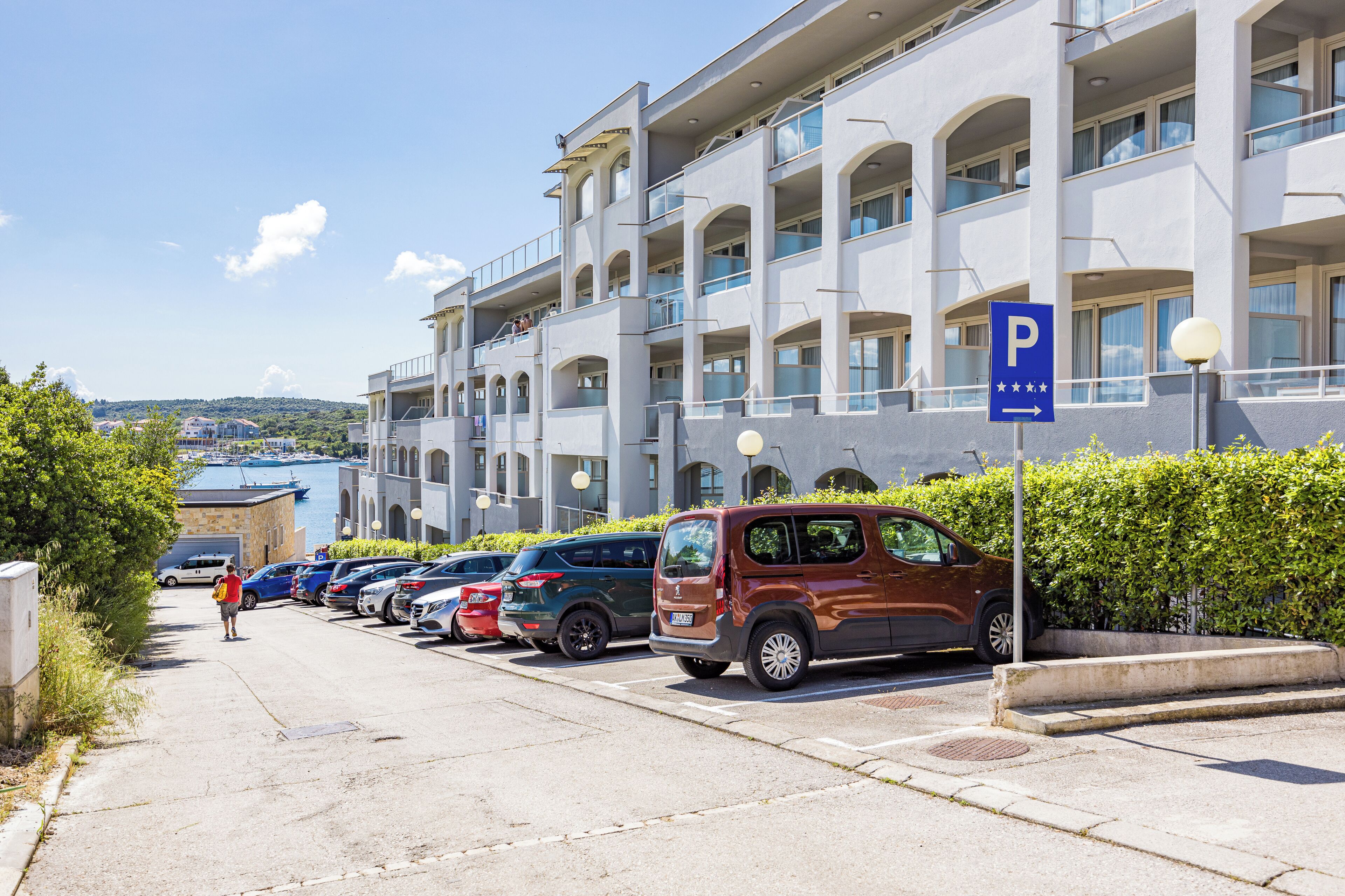 Ein Parkplatz mit einer Reihe geparkter Autos vor einem Gebäude mit Balkonen und Blick auf das Wasser.