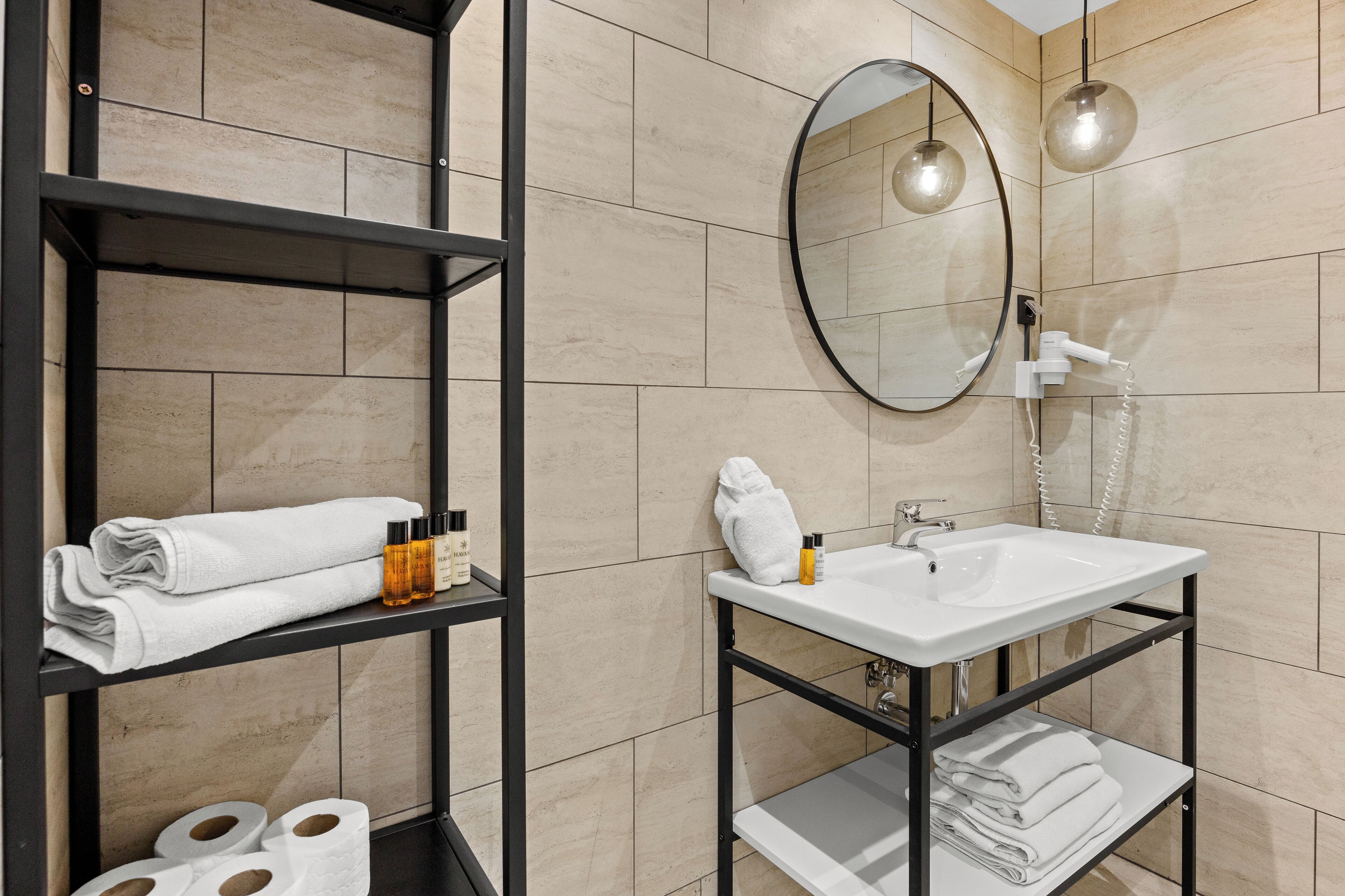 Ein Badezimmer mit weißem Waschbecken, einem Spiegel, Ablagen mit Handtüchern und Pflegeprodukten sowie einem Toilettenpapierhalter.