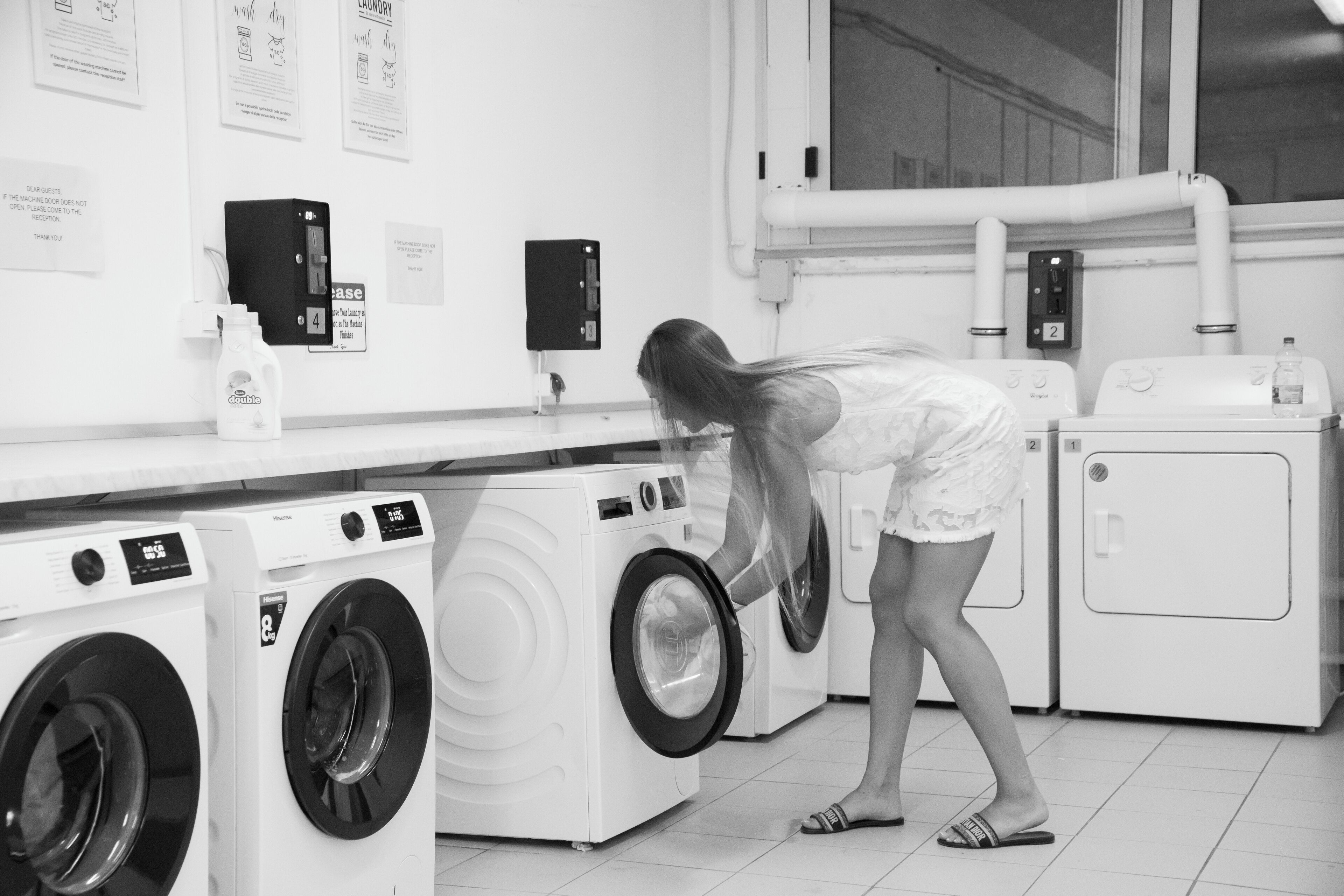 A laundromat with multiple washing machines and dryers, a person bending over a machine, and instructional signs on the wall.