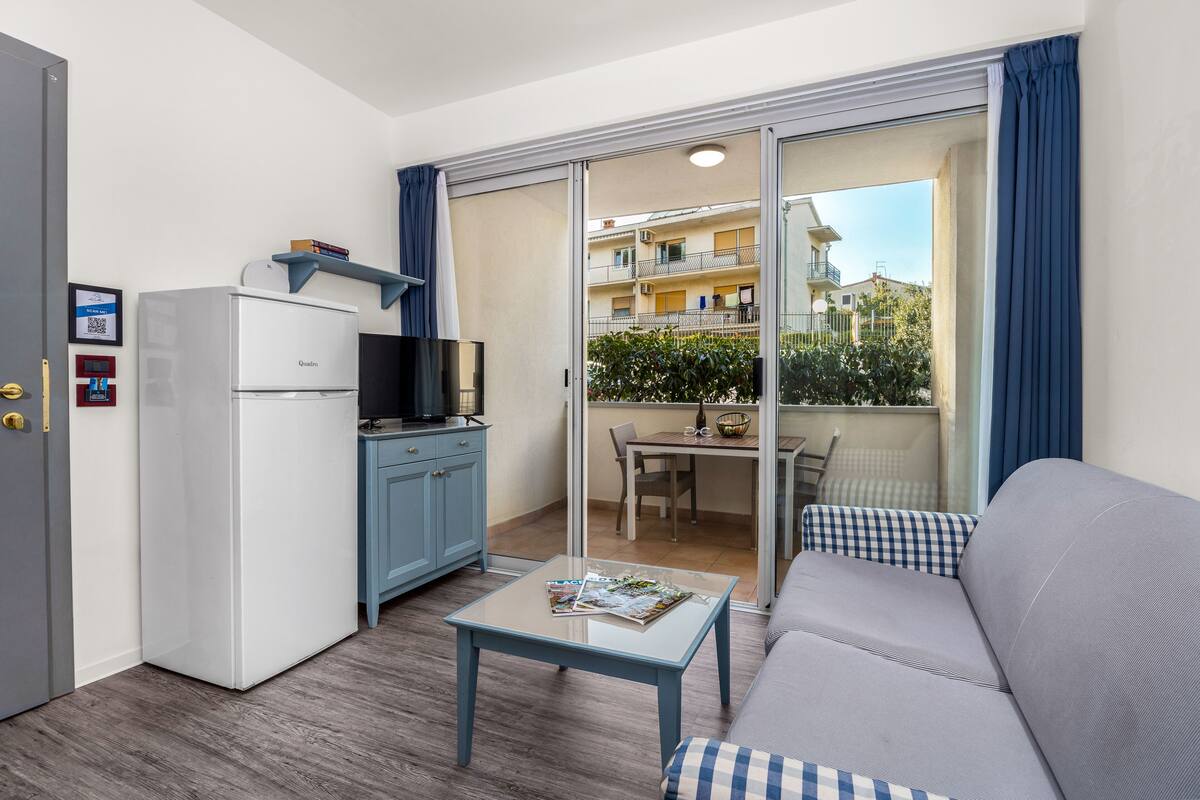 Un espacio habitable compacto con kitchenette, un sofá, una pequeña mesa y un balcón con vistas a los edificios circundantes.