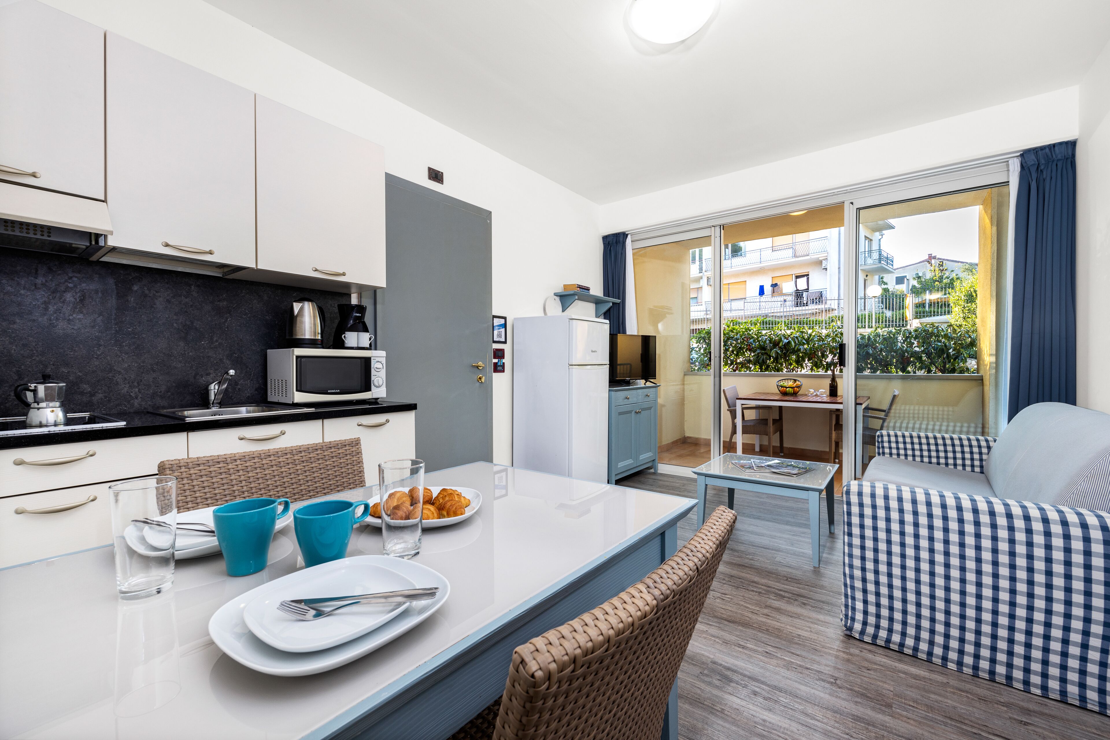 Een moderne keuken met witte kasten, een magnetron en een koffiezetapparaat. Er is een eethoek met een tafel, stoelen en uitzicht op het balkon.