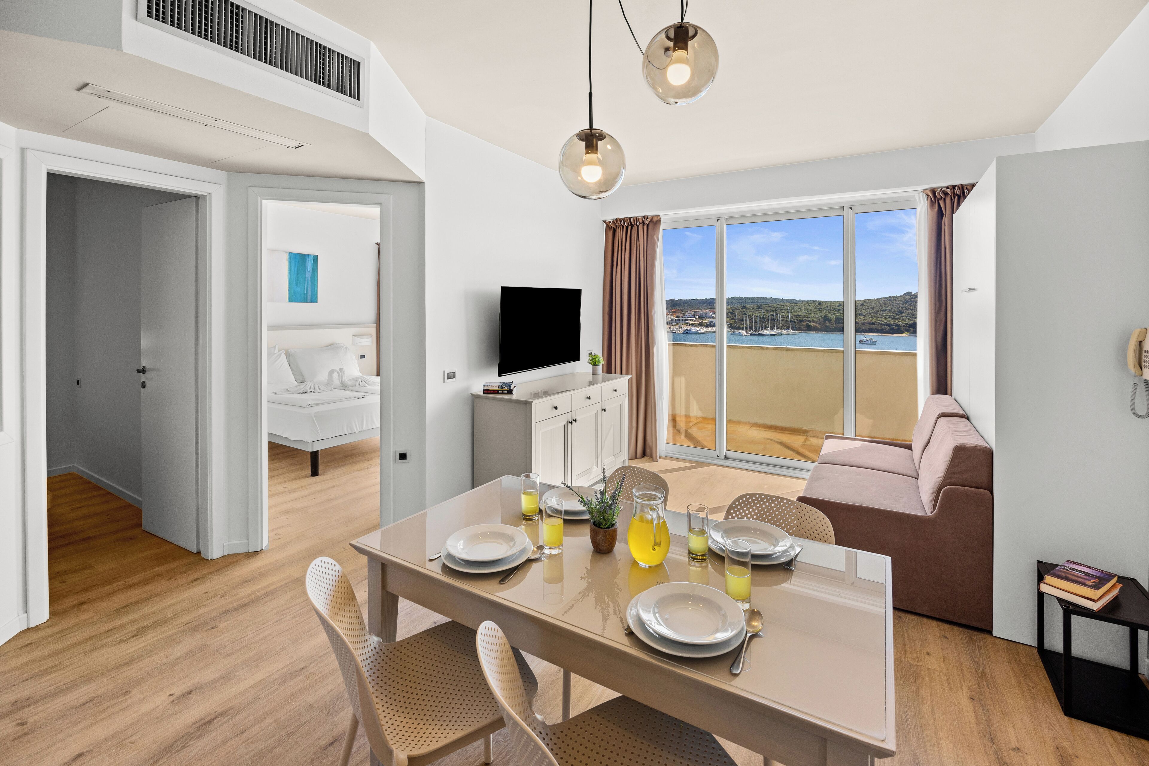 A modern room with a dining table set for four, a sofa, a TV, and a large window with a view of the sea.