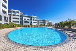A large, clear swimming pool surrounded by a tiled deck, with modern multi-story buildings in the background.